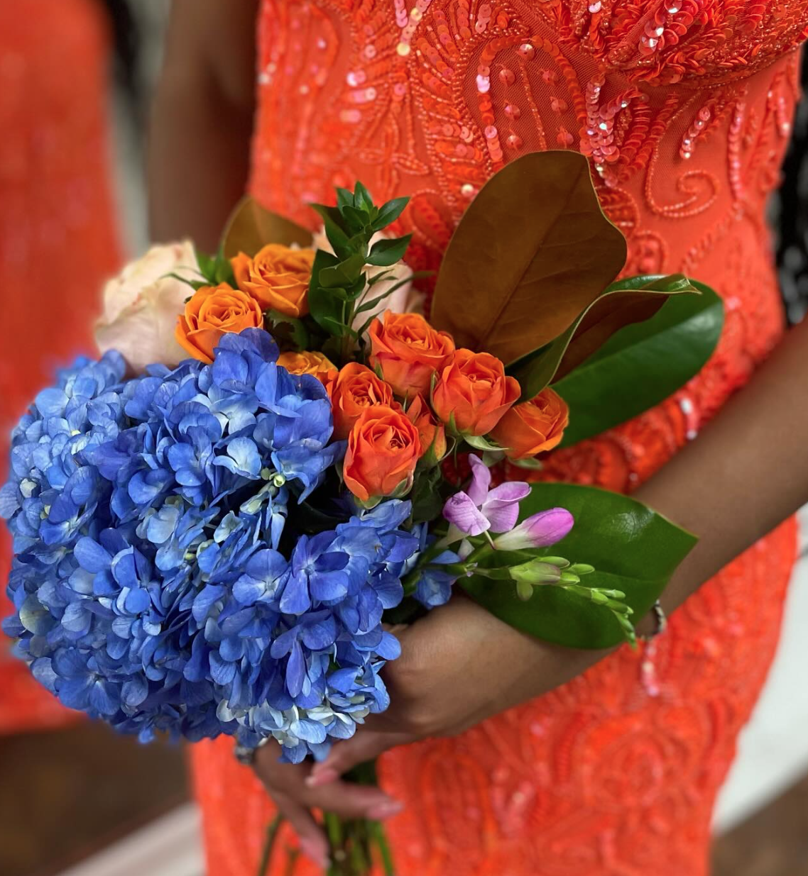 Person wearing a bright orange embroidered dress holding a bouquet of blue hydrangeas, orange roses, and pink buds, with green and brown leaves.