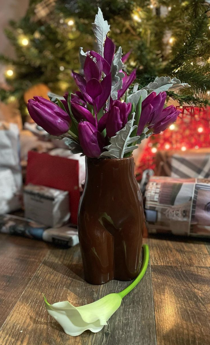 Purple tulips in a brown vase with a white calla lily flower lying on a wooden table, holiday decorations and a Christmas tree in the background.