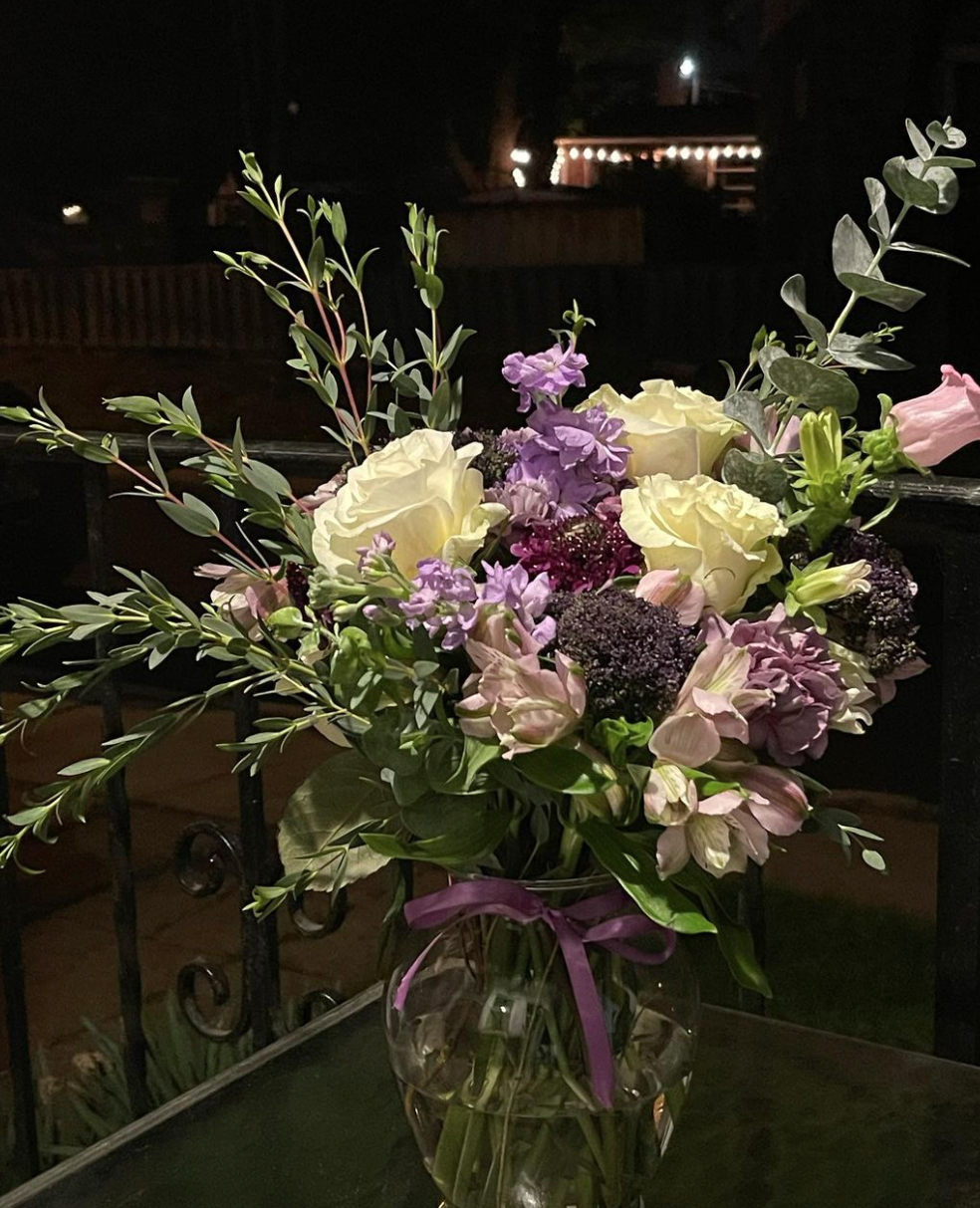An arrangement of white, purple, and pink flowers, including roses and other blooms, in a glass vase with a purple ribbon, set against a dark outdoor background at night.