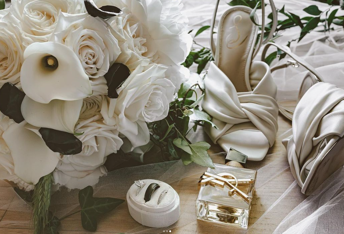 A close-up of a wedding bouquet of white roses, calla lilies, and greenery, along with various white and silver jewelry, a glass perfume bottle, white heels, and a white purse on a wooden surface with white fabric and tulle.