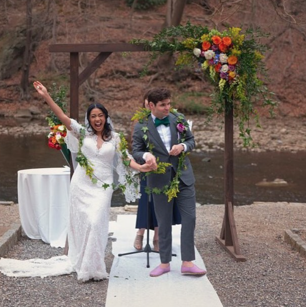 A bride and groom holding hands and celebrating at an outdoor wedding ceremony near a river, with a floral arch and colorful flower arrangements.