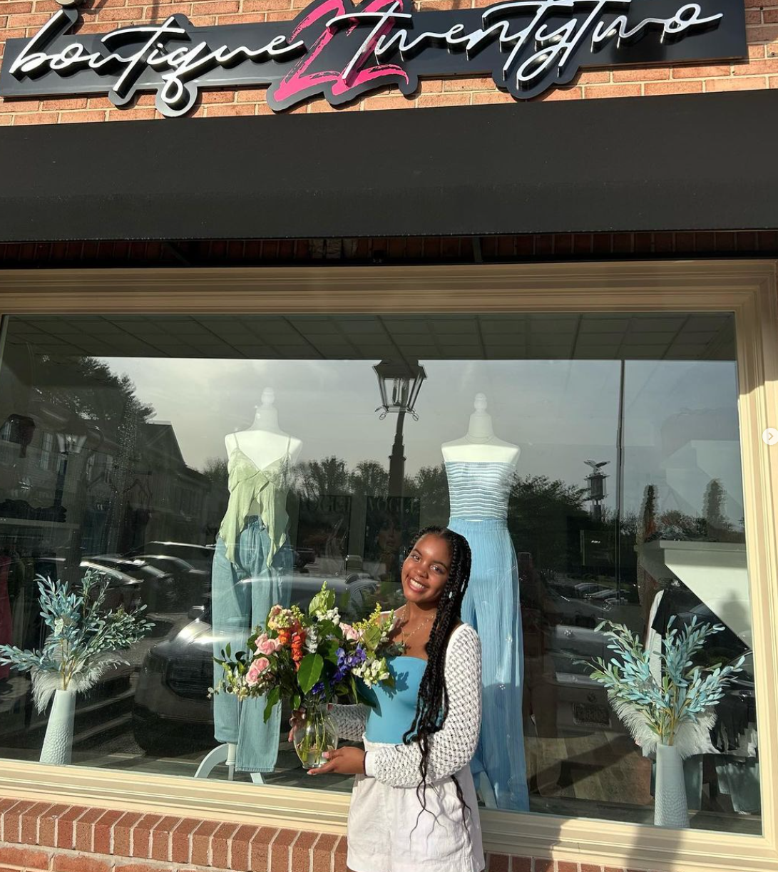 A smiling woman holding a large bouquet of flowers standing in front of a clothing store window display with mannequins dressed in summer outfits. The store sign above the window reads "boutique Tinasayo." The window reflection shows parked cars and 