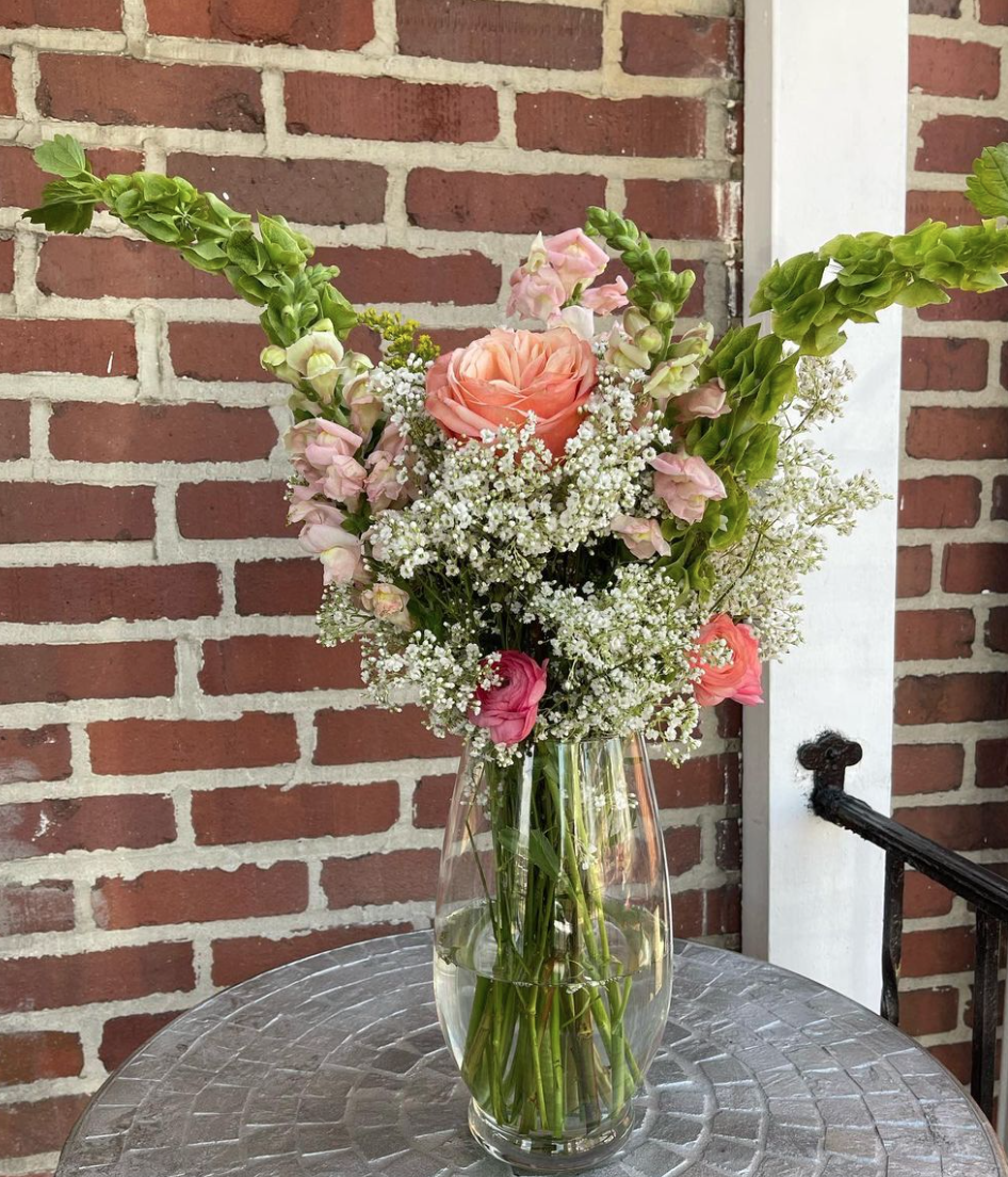 Pink and white flowers arranged in a clear glass vase on a metal table against a brick wall background.