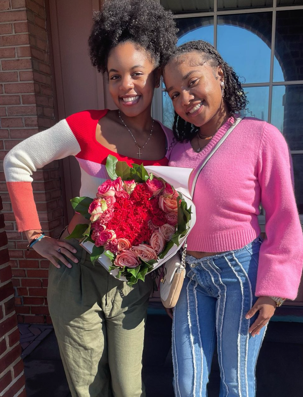 Two women standing outside in front of a brick building, smiling at the camera. One woman is holding a large bouquet of pink and red flowers, dressed in a colorful sweater and khaki pants, with her hair styled in a natural curly ponytail. The other w