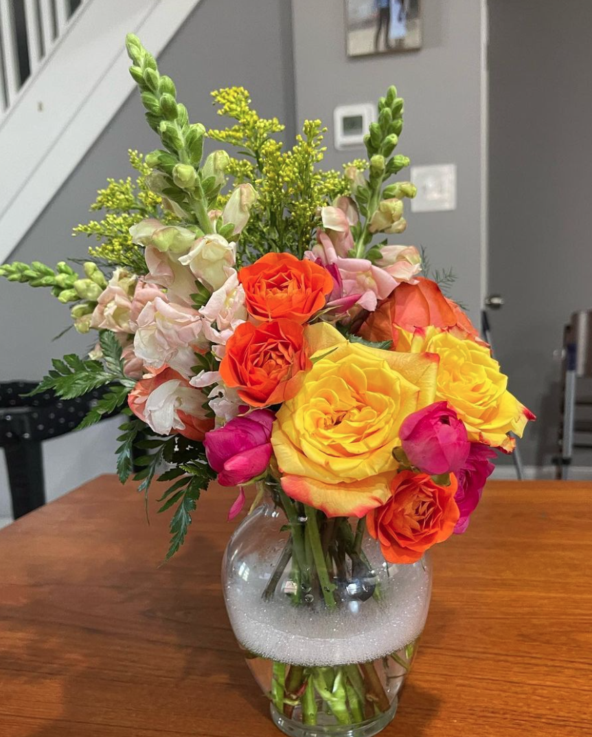 A colorful bouquet of flowers in a clear glass vase on a wooden table. The bouquet includes yellow roses, orange roses, pink roses, white snapdragons, and green foliage.