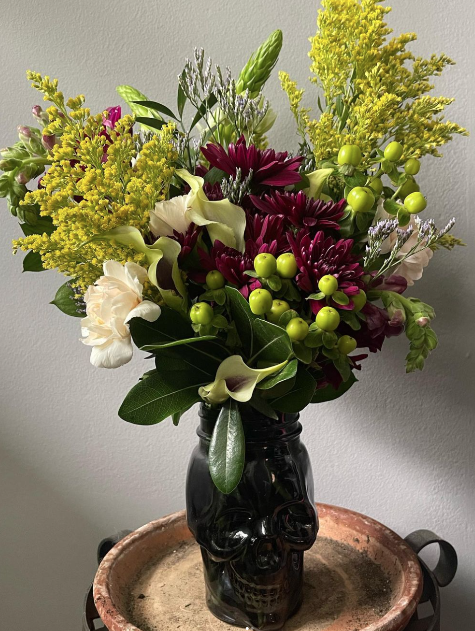 A floral arrangement in a black skull-shaped vase featuring yellow, purple, and white flowers with green foliage, placed on a round table against a plain wall.