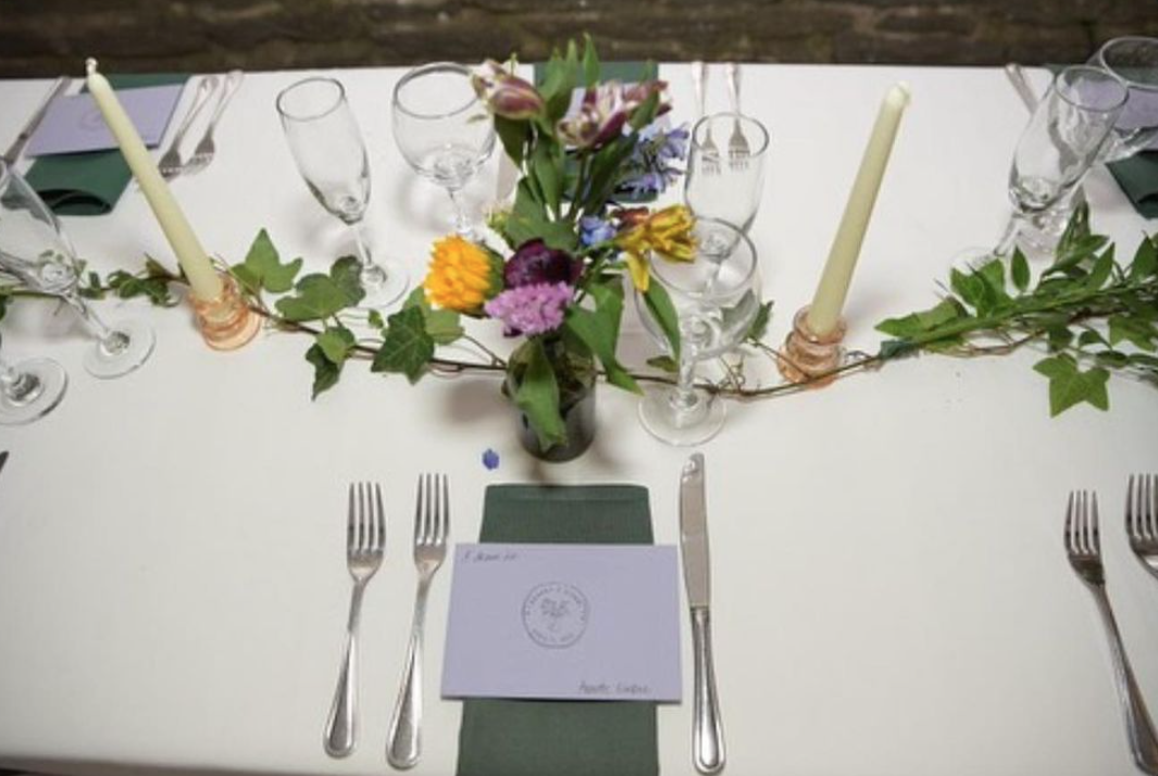 Table setting with white tablecloth, green napkin, silverware, clear wine glasses, a paper menu, candles, and a floral centerpiece with various colorful flowers and greenery.