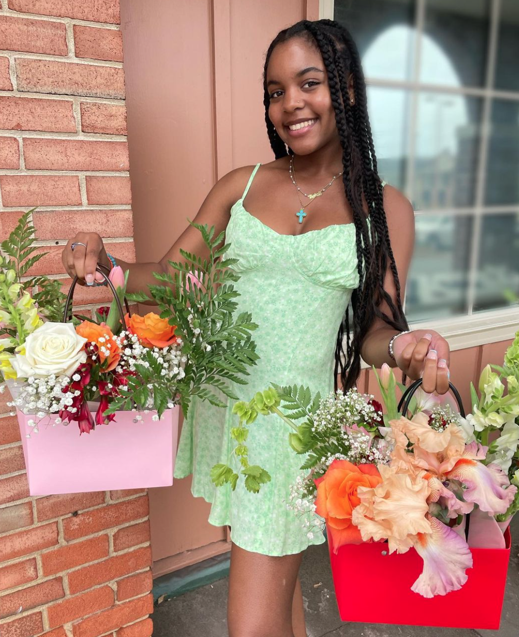 A young woman with braided hair, wearing a light green dress, smiling while holding two flower bouquets in pink and red baskets outside a brick building.