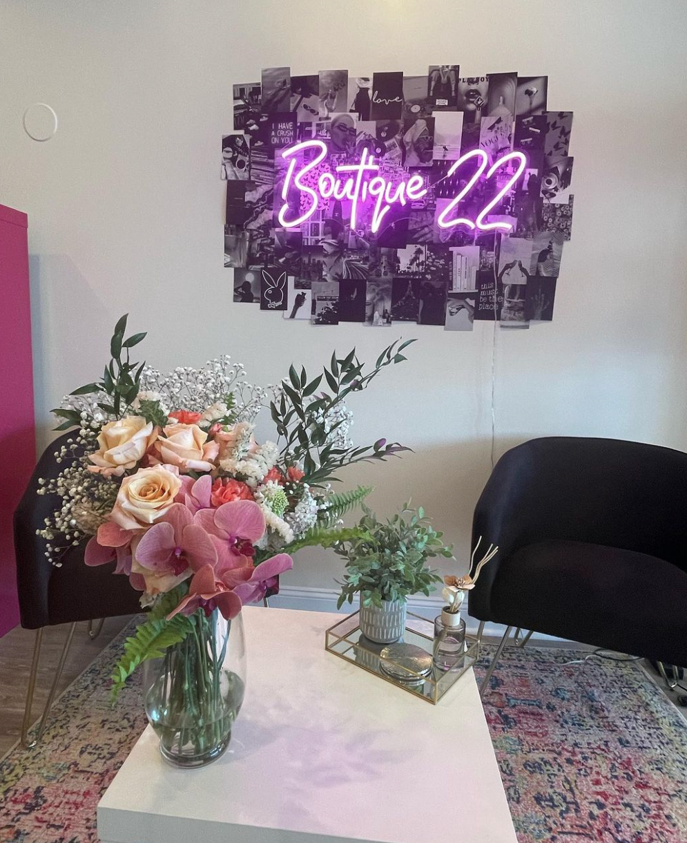 Interior scene with a white table holding a large floral arrangement, and a wall decorated with a neon sign that reads 'Boutique 22' in pink, surrounded by a collage of black-and-white photos, with two black chairs nearby.