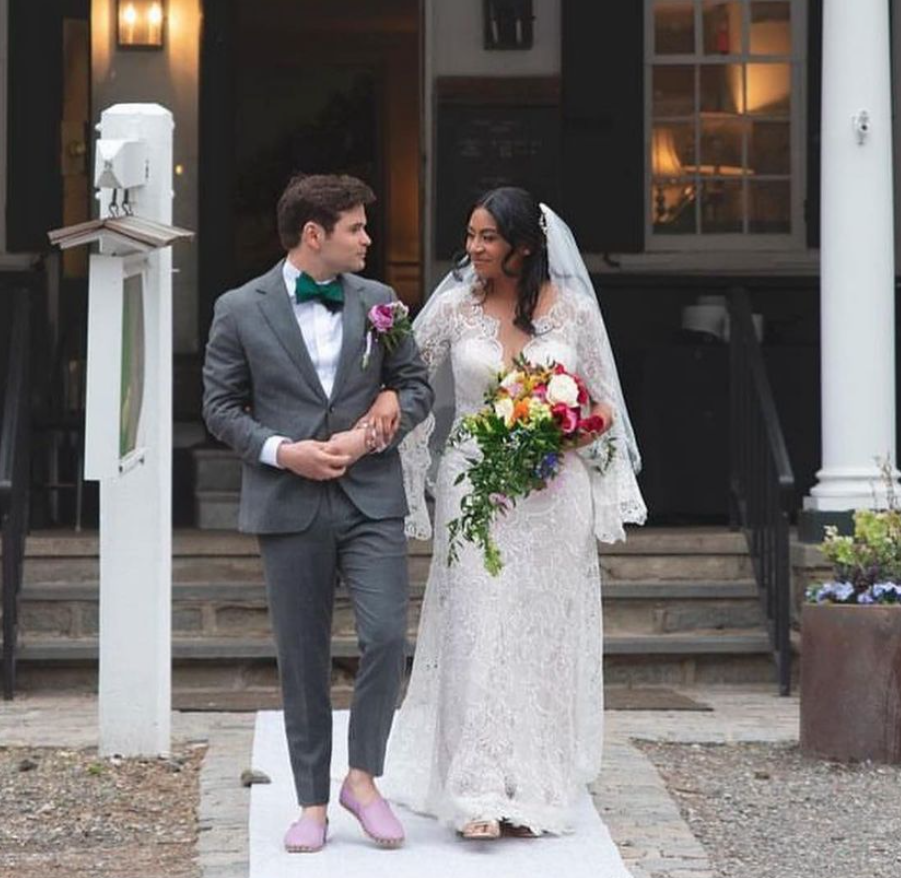 Couple dressed in wedding attire walking down the aisle outside a house, with the bride holding a colorful bouquet of flowers.