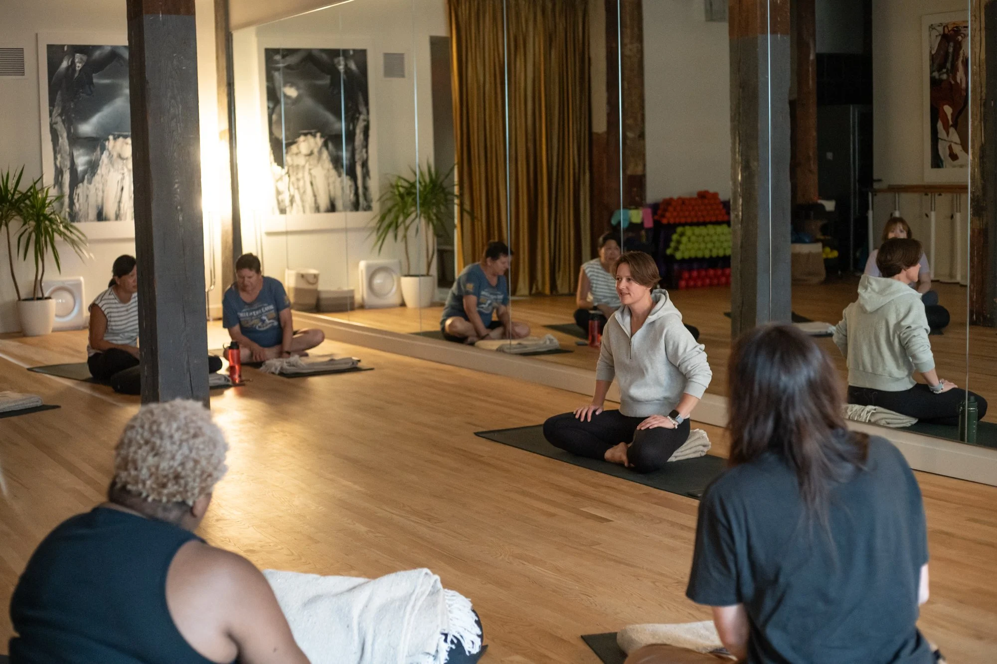 People participating in a yoga class in a studio with wooden floors, taped mats, and large mirrors, with exercise equipment and abstract paintings on the walls.