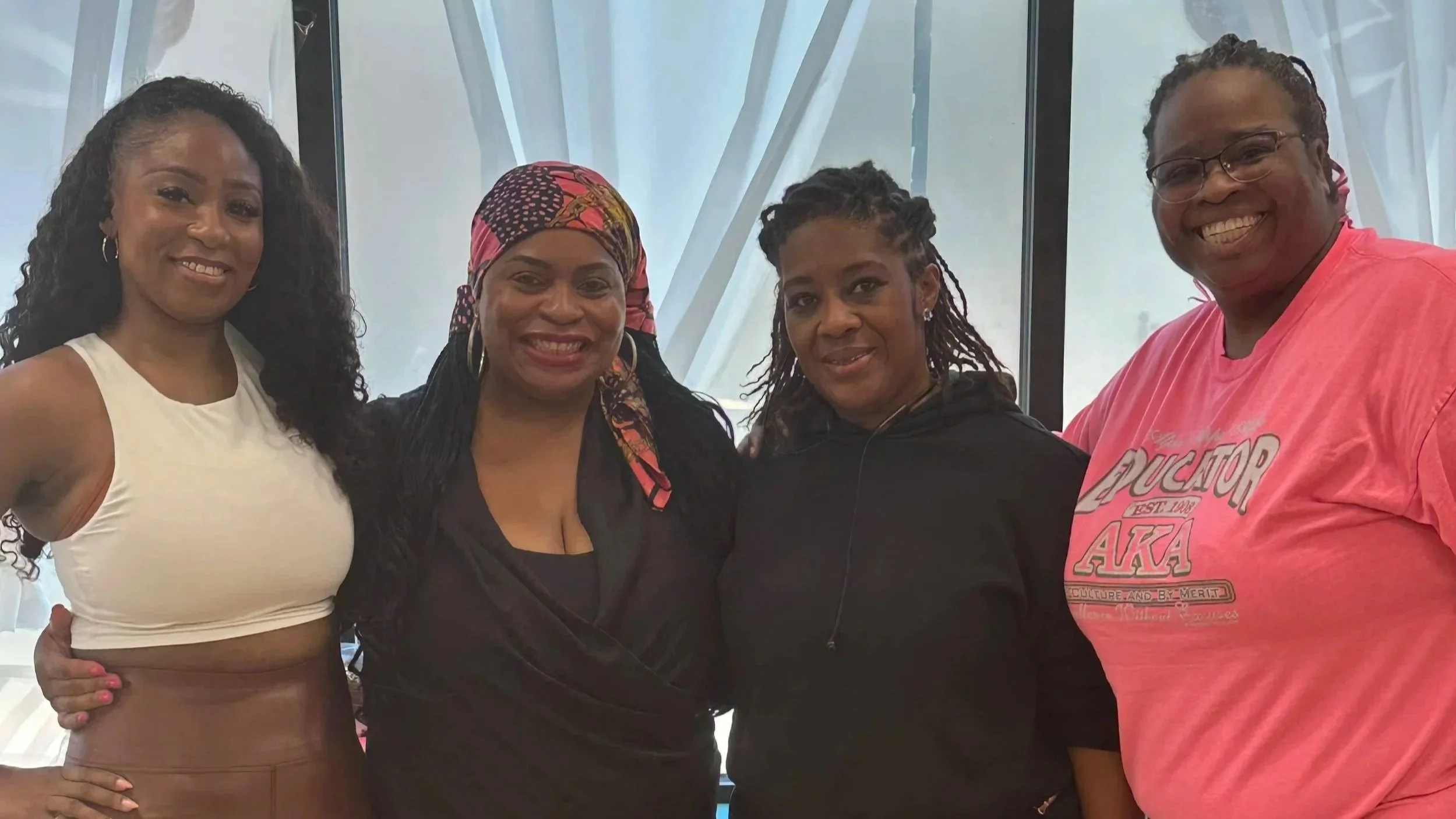 Four women standing close together, smiling, in front of a window with white curtains.