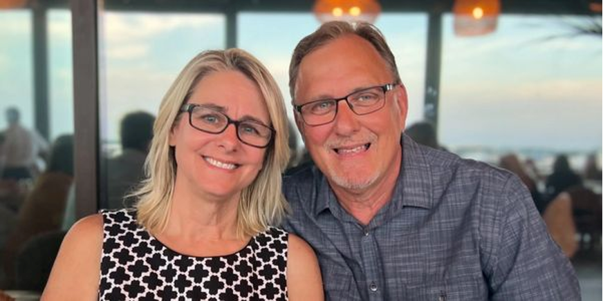 Pastor Jon Stoe and his wife Tedi smiling and sitting together indoors with a window view of the ocean and a sunset in the background.