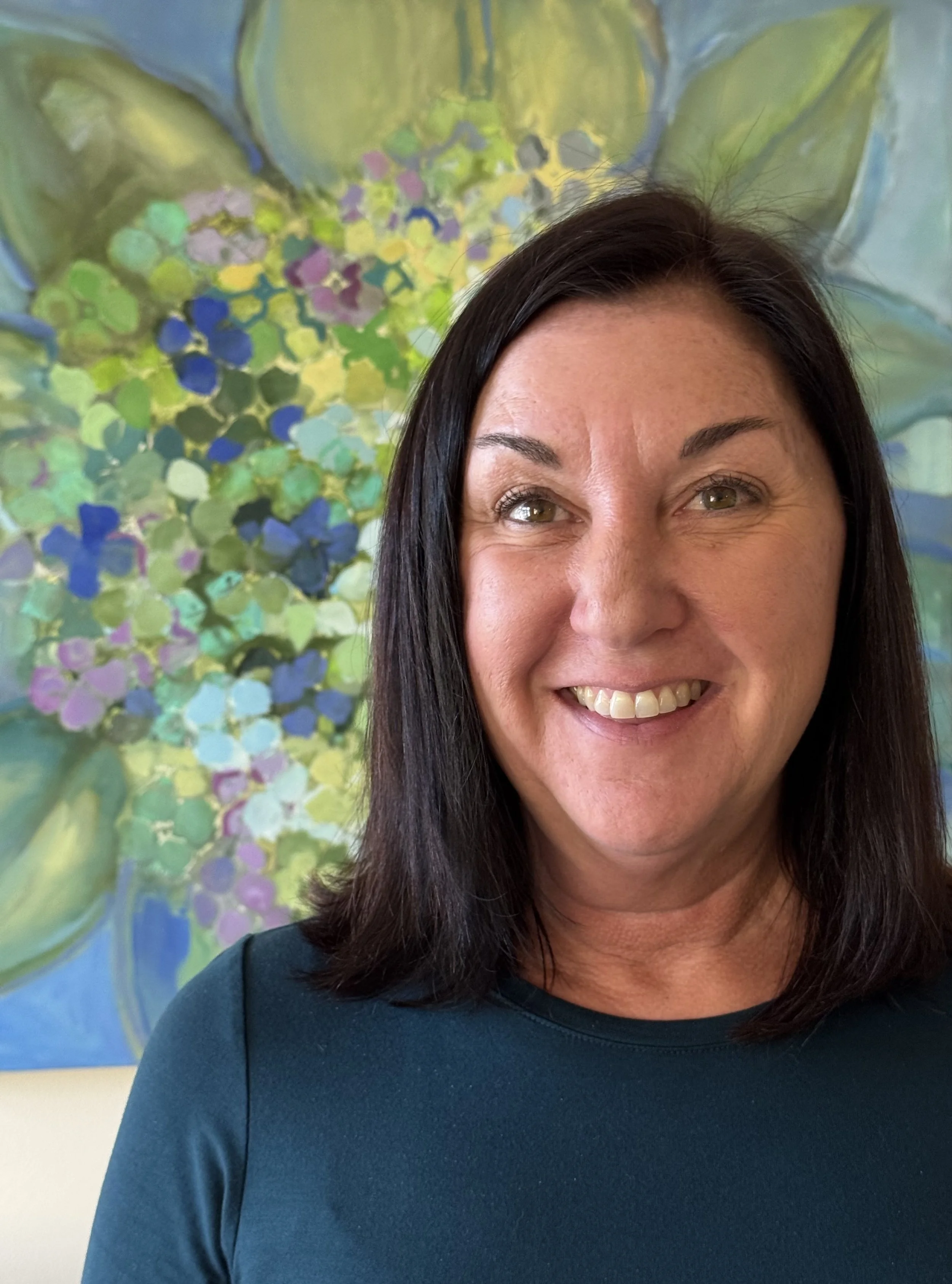 Smiling woman with long brown wavy hair sitting on a dark blue couch with white faux fur pillow in a light-colored room.