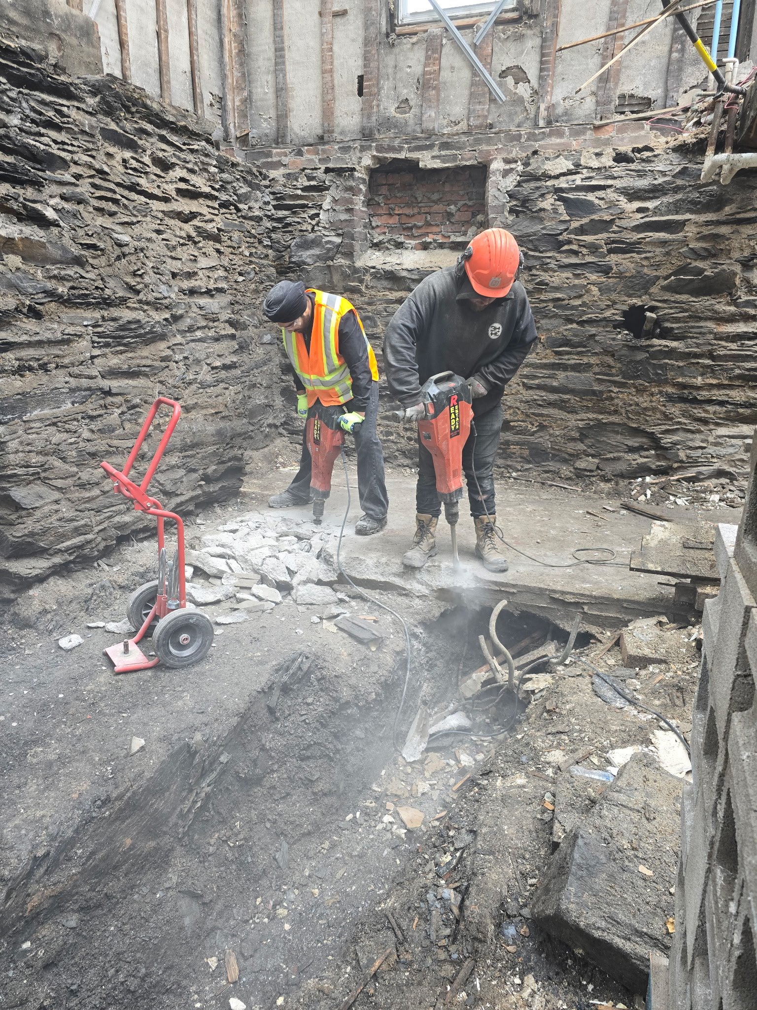 Two construction workers underground using jackhammers on the ground, surrounded by dirt and debris, with exposed brick and concrete walls behind them.