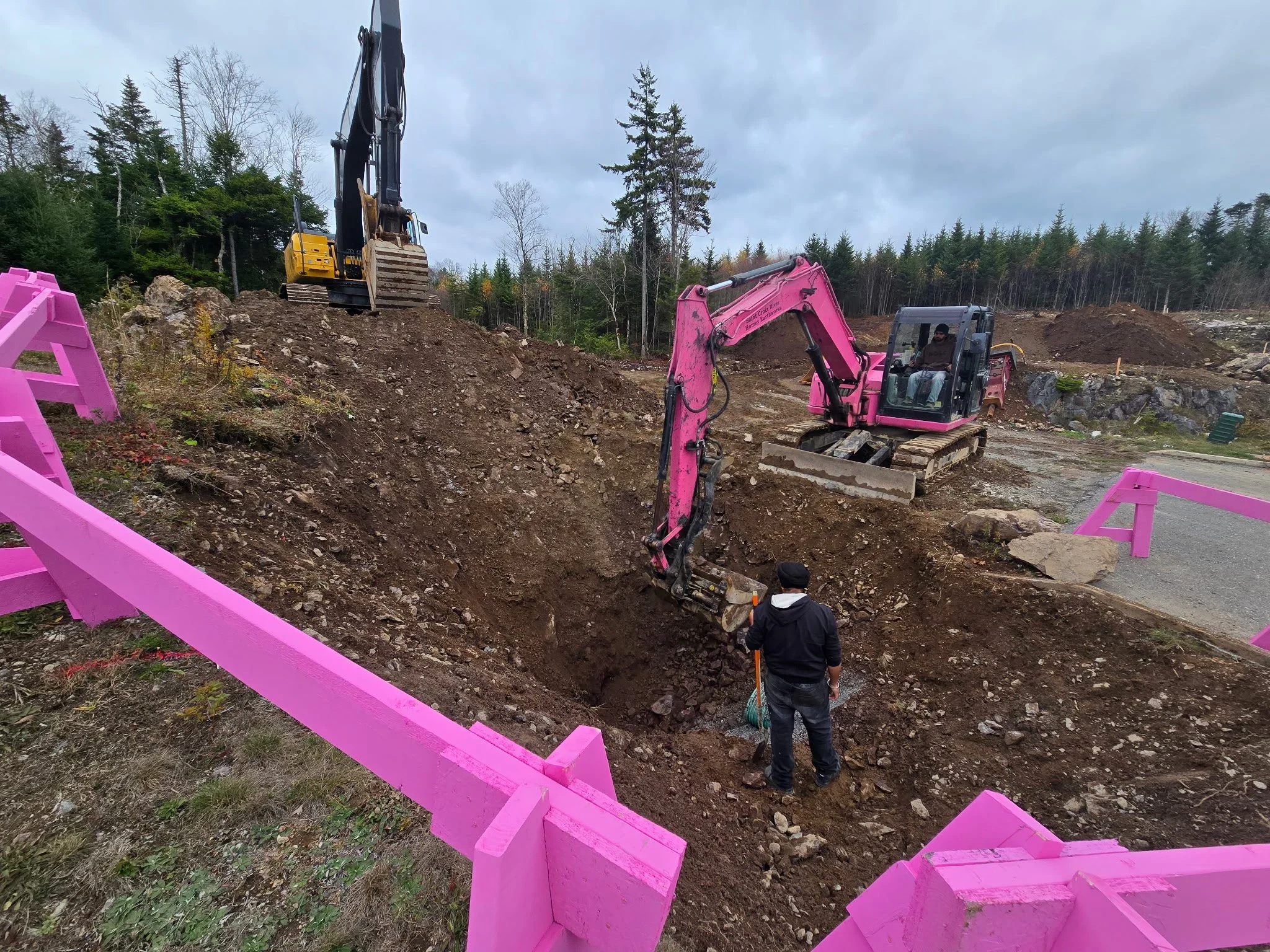 Construction site with two excavators working on a slope; one excavator is pink and the other is yellow; pink safety barriers are around the site; two workers are present, one operating the pink excavator and another standing in the dirt with a broom; background features trees and a cloudy sky.