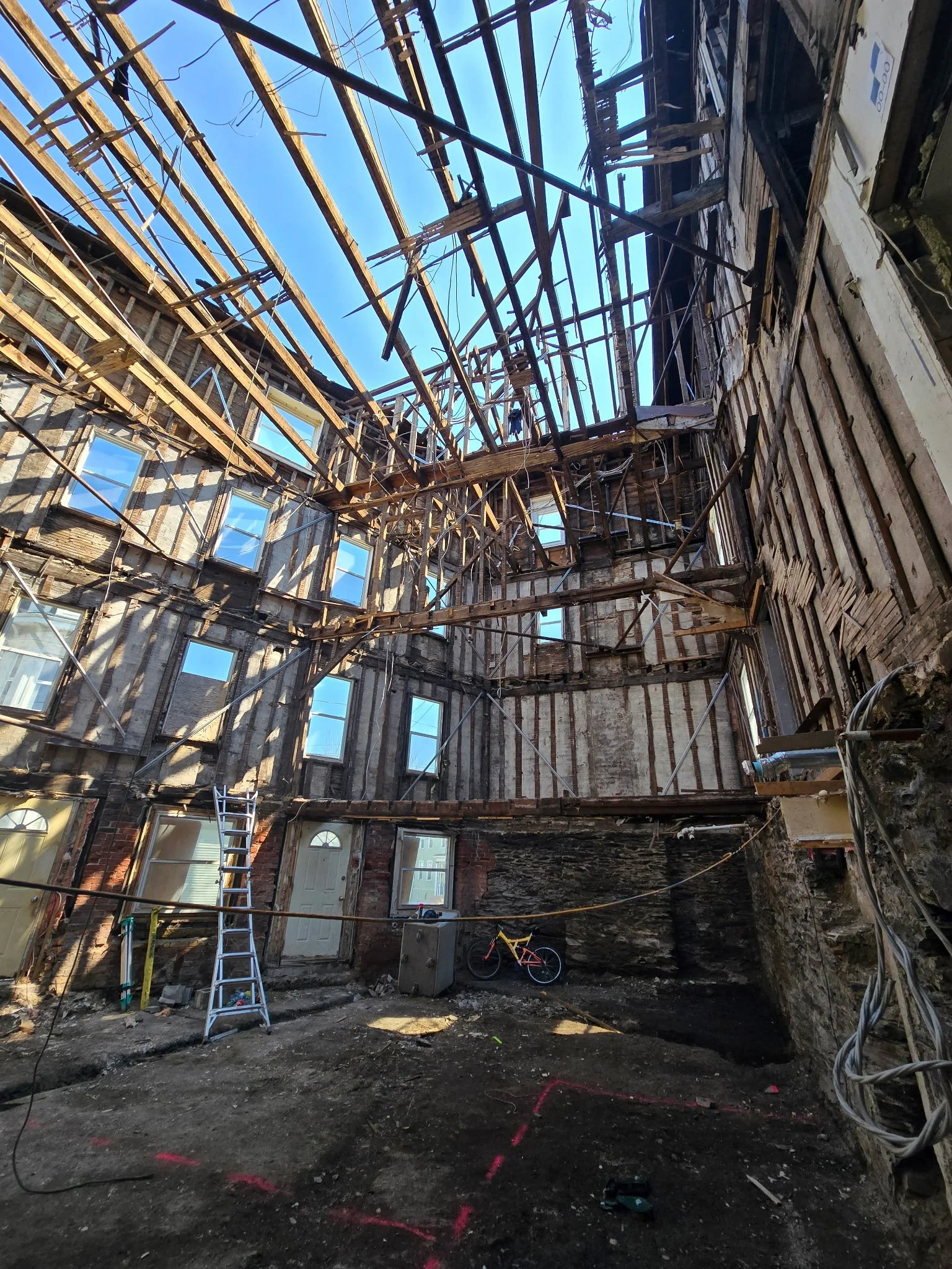 The interior of a building under renovation with exposed wooden beams, scaffolding, and construction debris on the ground, with a blue sky visible through the roof opening.