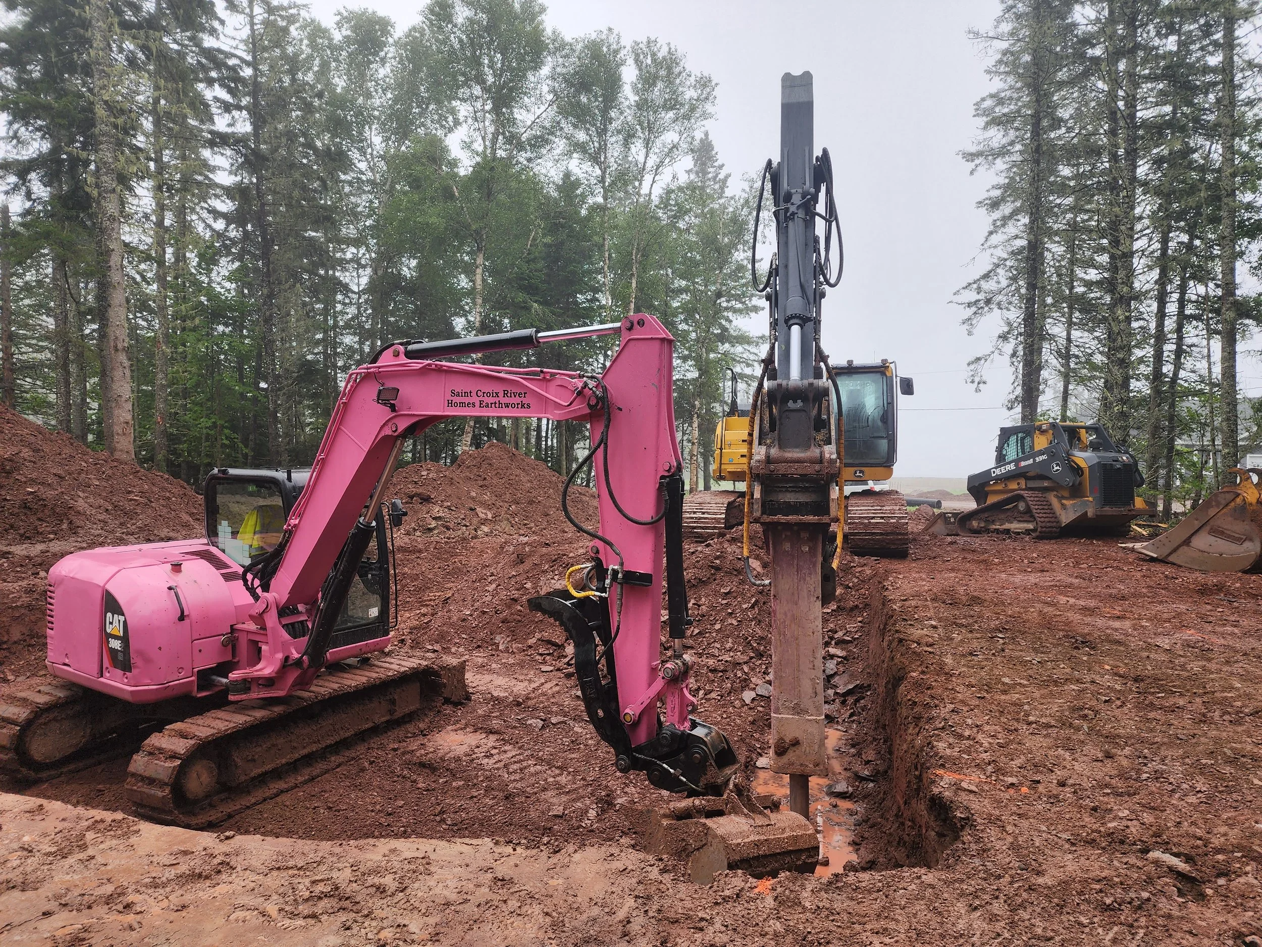 A construction site with a pink excavator, and additional yellow and black heavy machinery in a wooded area, with cleared dirt and trees in the background.