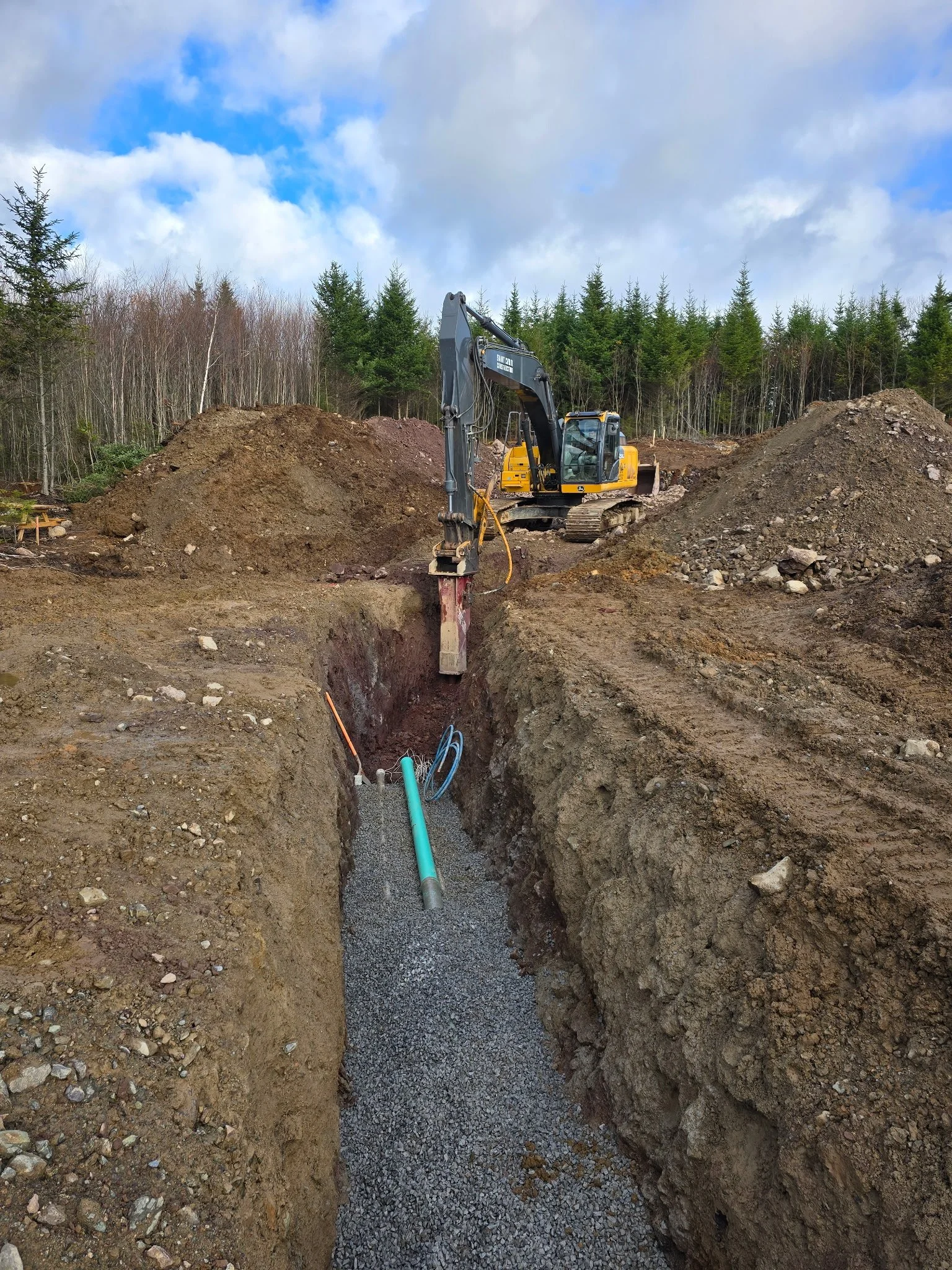 Construction site with a yellow excavator digging a trench, with soil piles on either side and underground pipes and gravel inside the trench, against a backdrop of trees and cloudy sky.