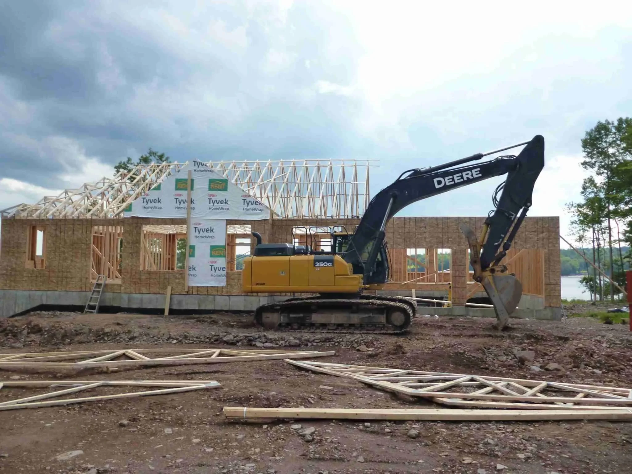 Construction site with a yellow Deere excavator working on a wooden frame house under a cloudy sky, with building materials and trees nearby.