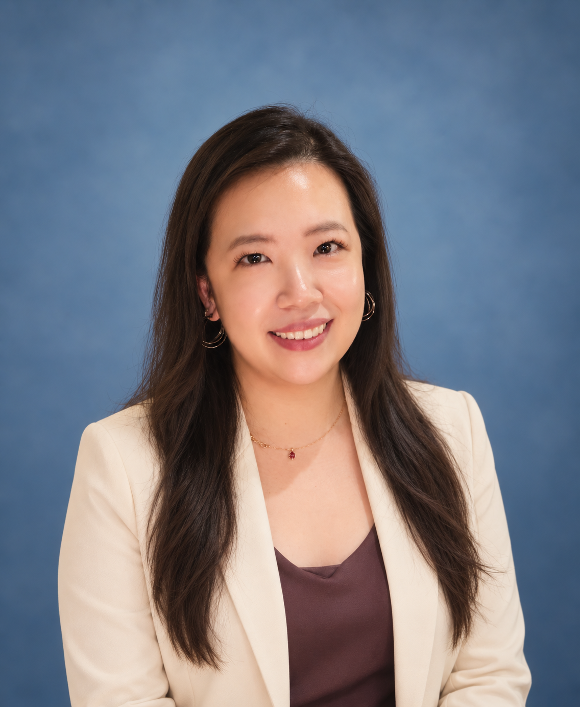 Professional headshot of a smiling female therapist wearing a cream blazer against a soft, muted blue background.