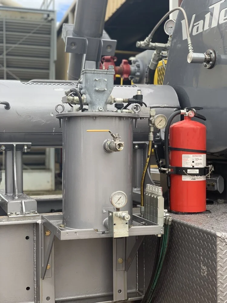 Close-up of industrial machinery with a gray cylindrical tank, gauges, pipes, and a red fire extinguisher in an outdoor setting.