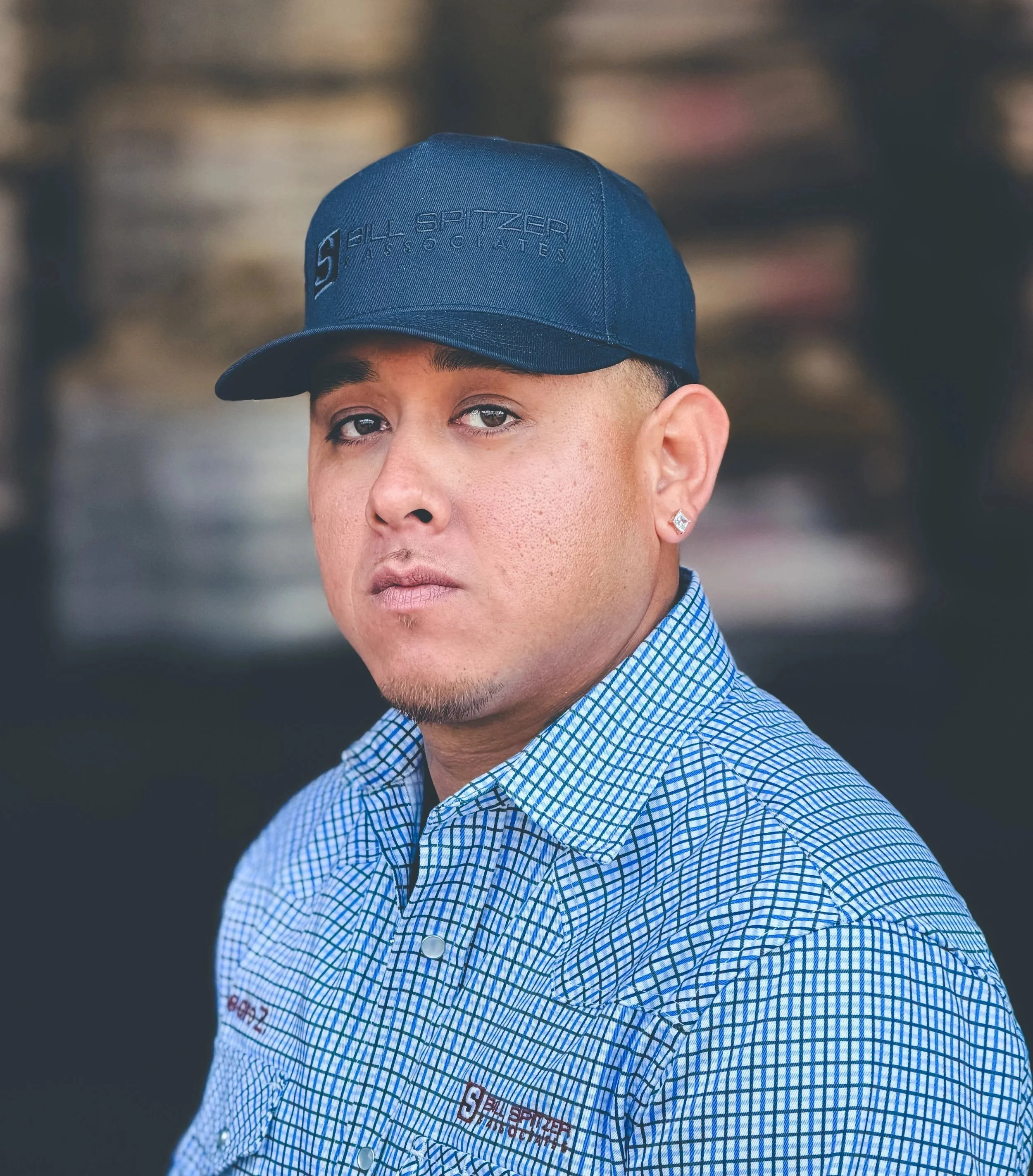A smiling man with a beard wearing a blue patterned shirt and a gray baseball cap with the logo 'Bill Spitzer & Associates' stands outdoors against a dark blurred background.
