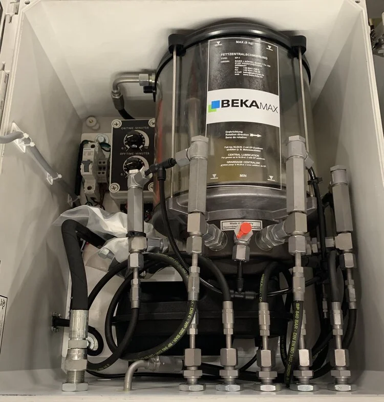Industrial or mechanical equipment with a large cylindrical component labeled BEKAMAX, various pipes, valves, and electrical connections arranged inside a white enclosure.