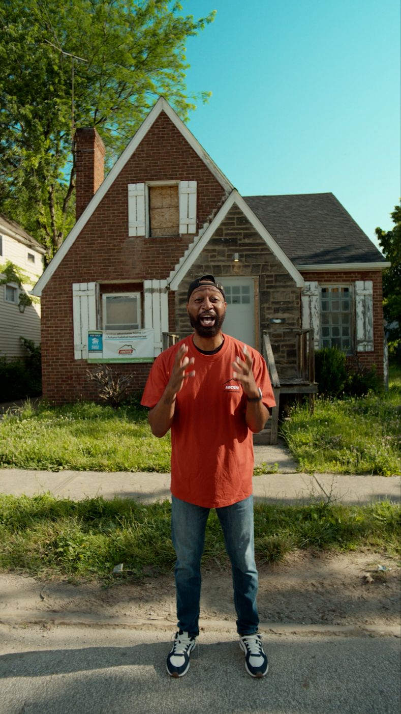 Man in red shirt standing in front of a house with boarded-up windows and a damaged exterior, speaking passionately.