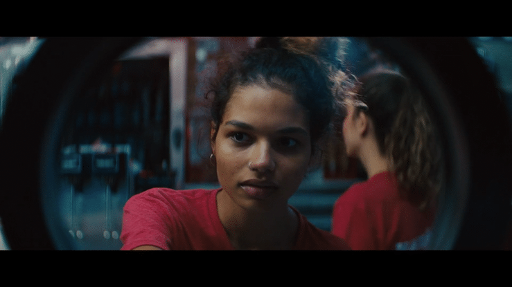 Young woman with curly hair in a red shirt looking into a circular mirror inside a laundry room.