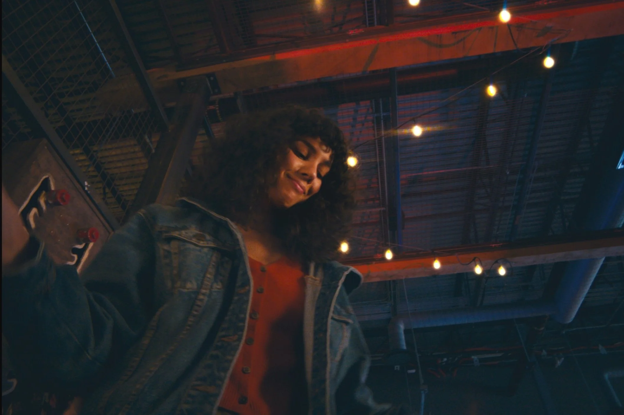 A woman with curly hair smiling while looking down, wearing a denim jacket and red shirt in a dimly lit venue with string lights overhead.