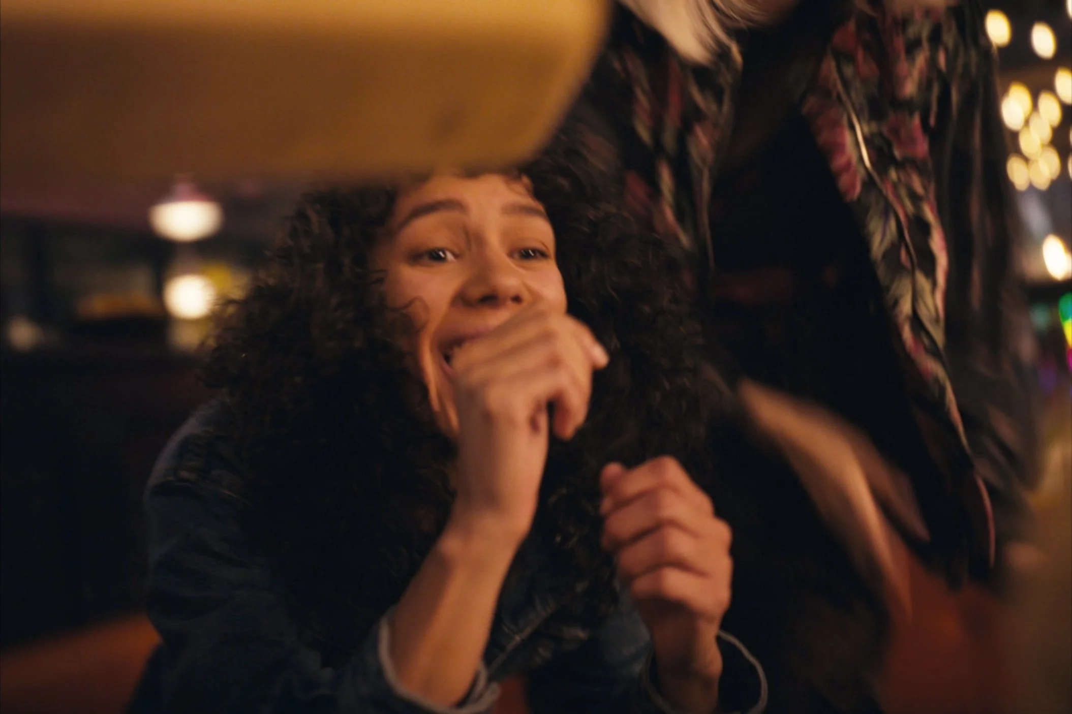 A woman with curly hair covering her mouth, appearing to be laughing or crying, in a dimly lit bar or club environment.