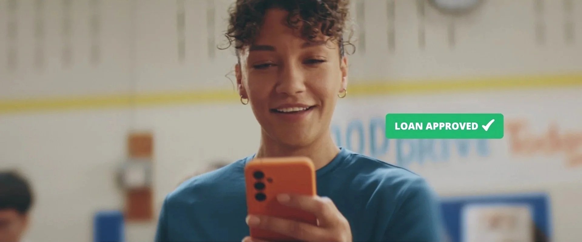 A young woman with short curly hair and earrings smiling while looking at her phone in a laundromat, with a green overlay text that says 'Loan Approved' and a checkmark.