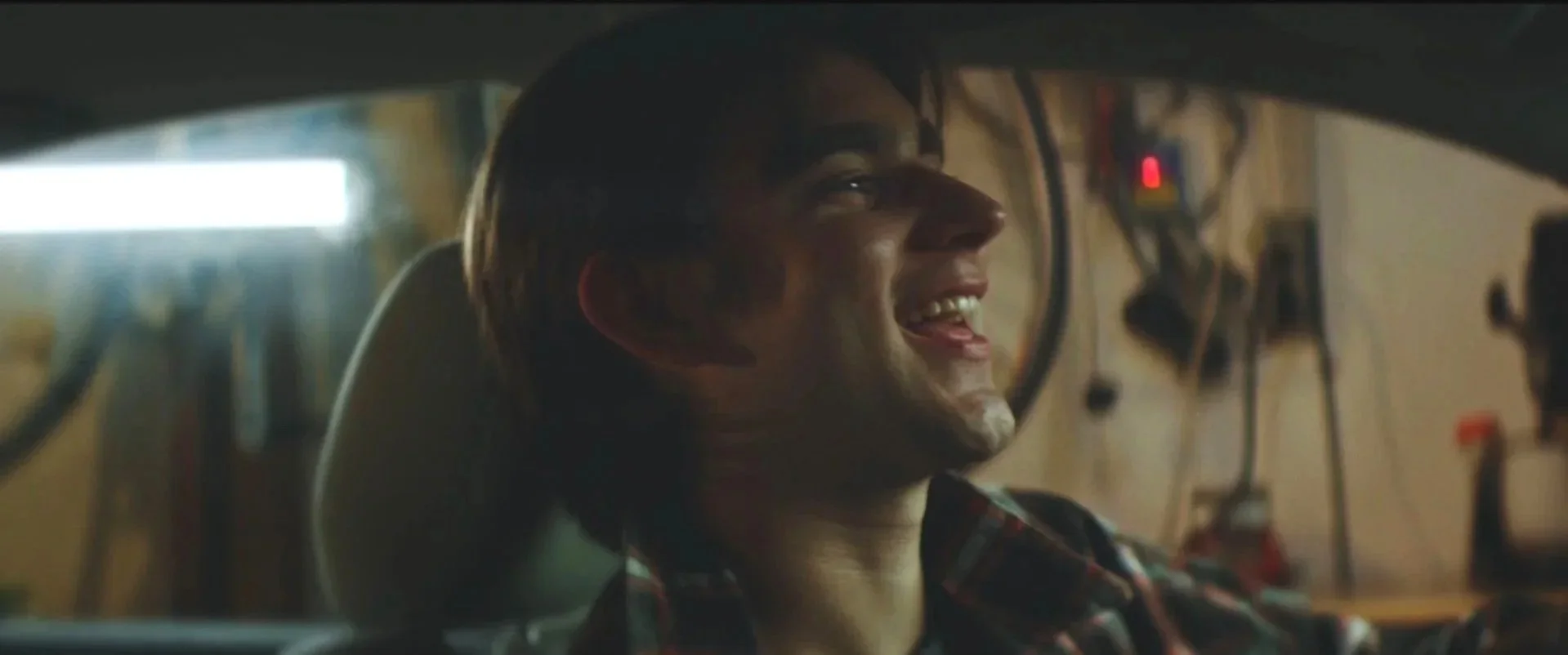 A young man with glasses smiling while sitting inside a car workshop.