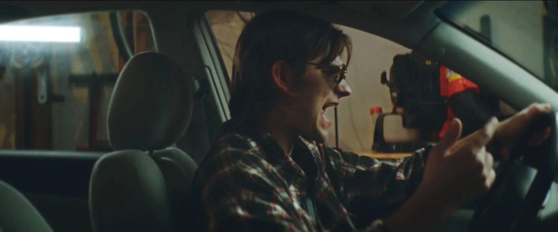A young man with dark hair and sunglasses sitting in a car, shouting or yelling, with a background of a garage or workshop.