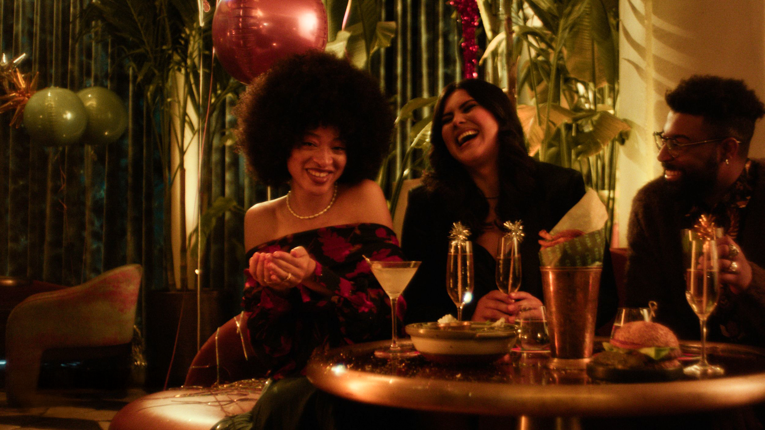 Three people, two women and a man, sitting at a table during a celebration, smiling and laughing, with balloons and festive decorations in the background.