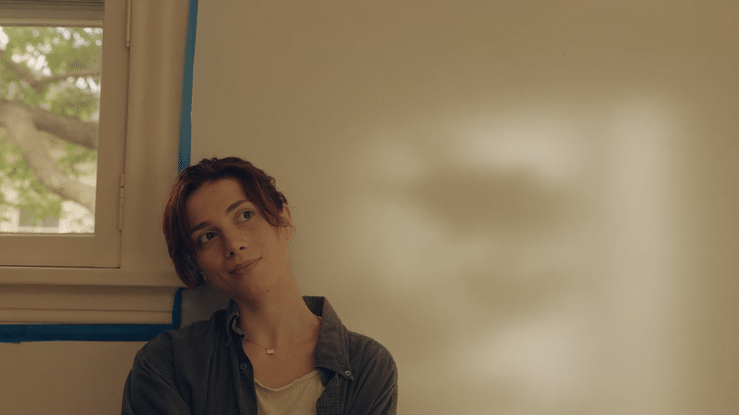 A woman with short red hair sitting in a room with beige walls and a window showing green trees outside.