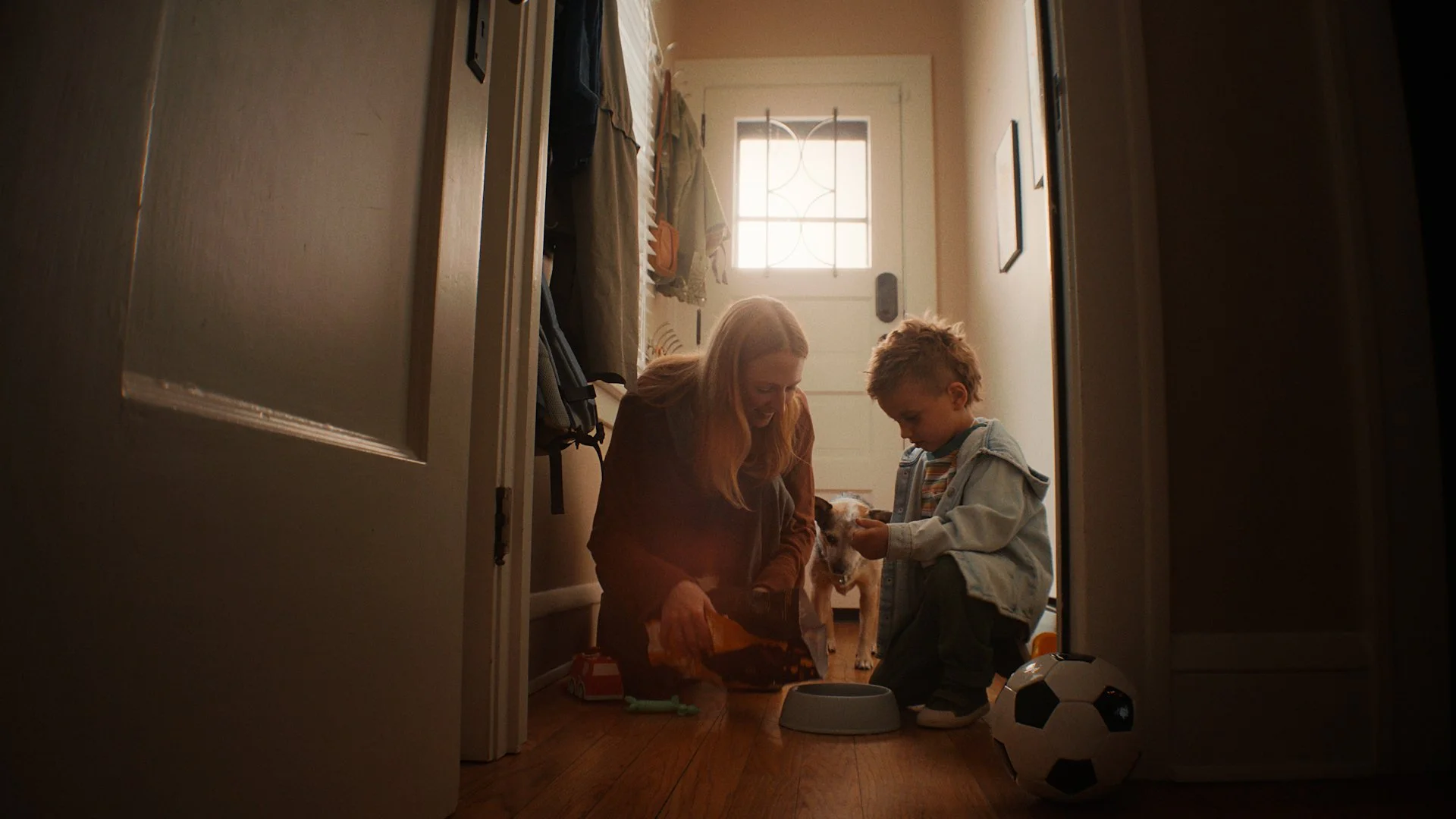 A woman and young boy sitting on the floor in an entryway, feeding a dog from a bowl. A soccer ball is nearby, and a backpack hangs on the wall.