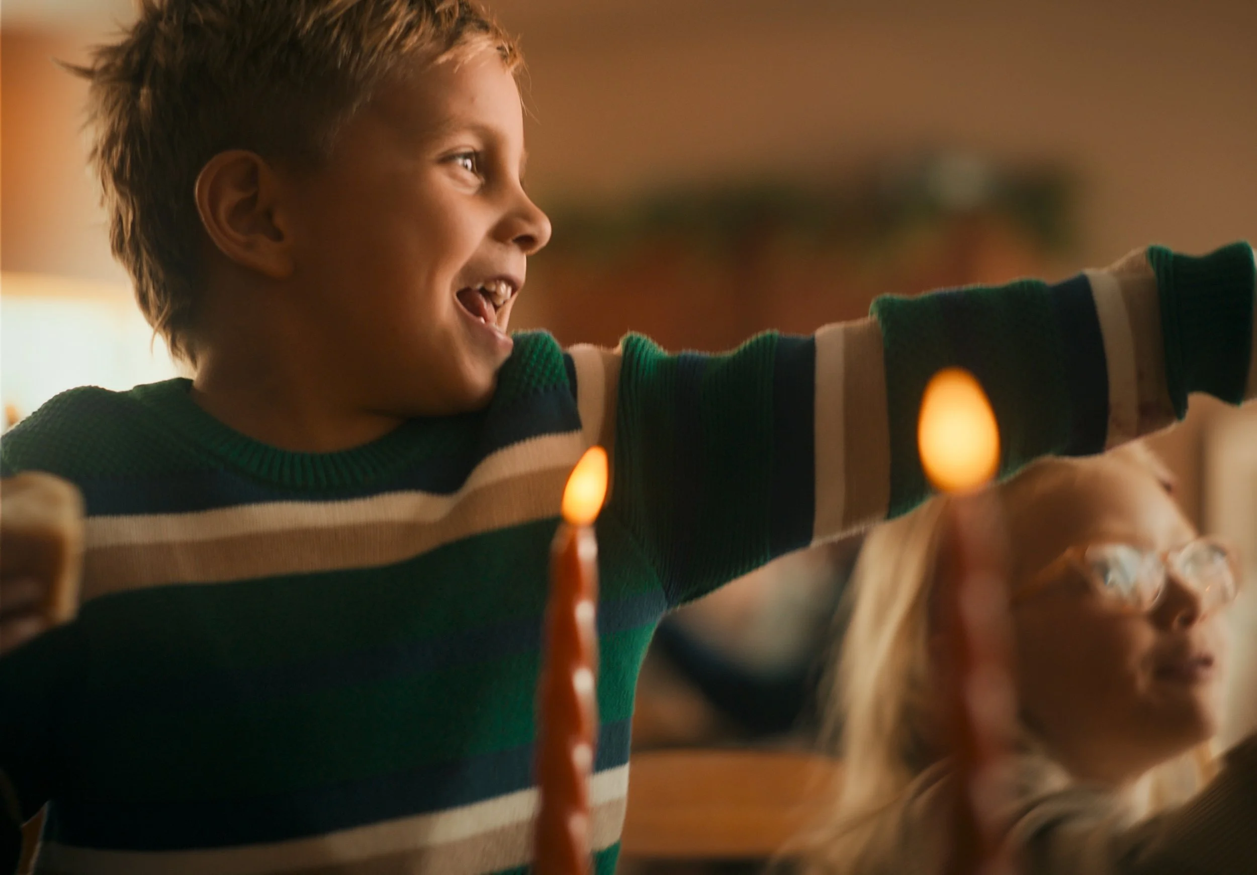 A young boy with short, spiky hair smiling and reaching out during a birthday celebration, with a lit birthday candle in the foreground and a girl with glasses in the background.