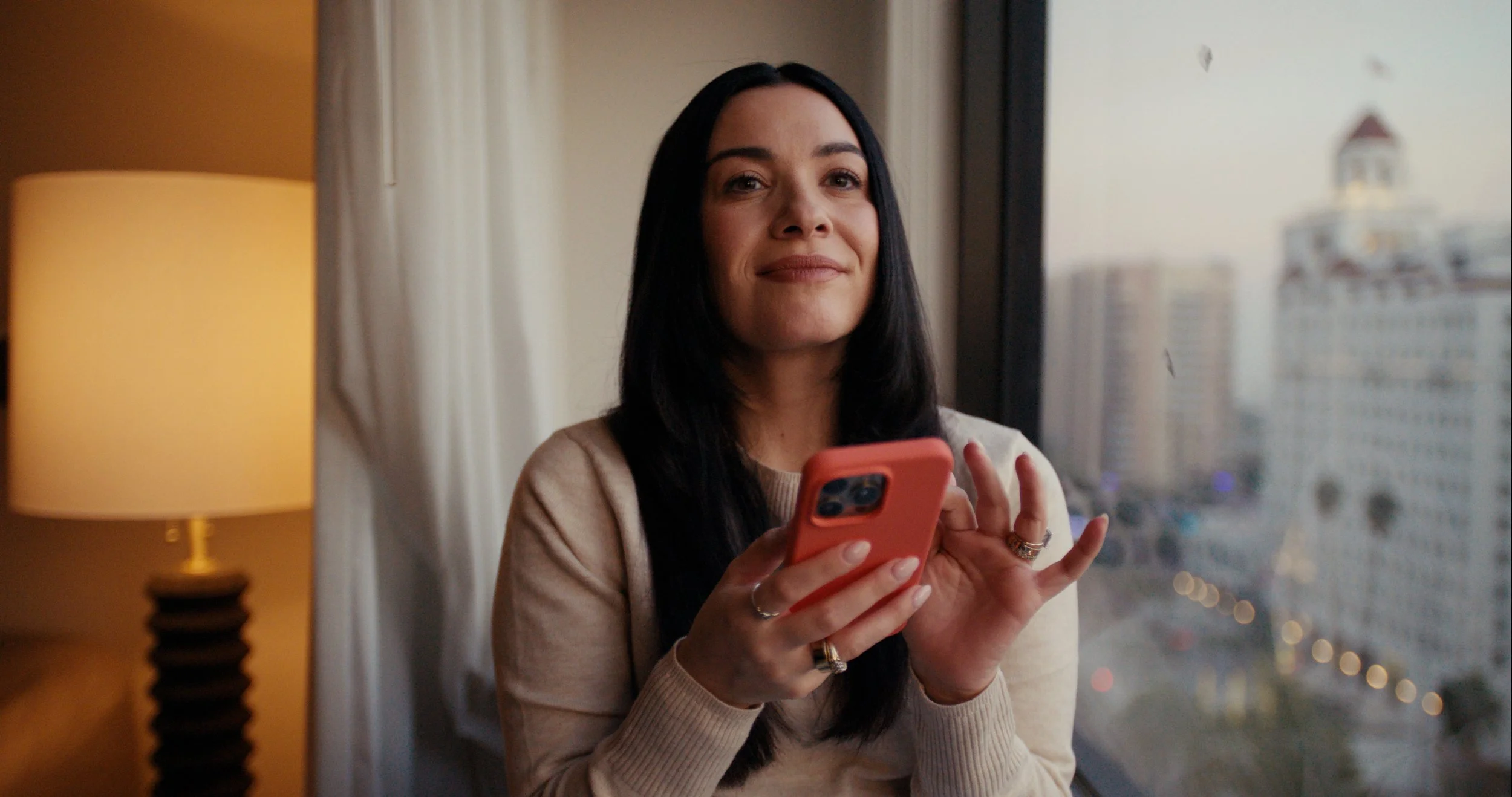 Woman with long black hair smiling and holding a pink smartphone while sitting by a window in a hotel room with city view.
