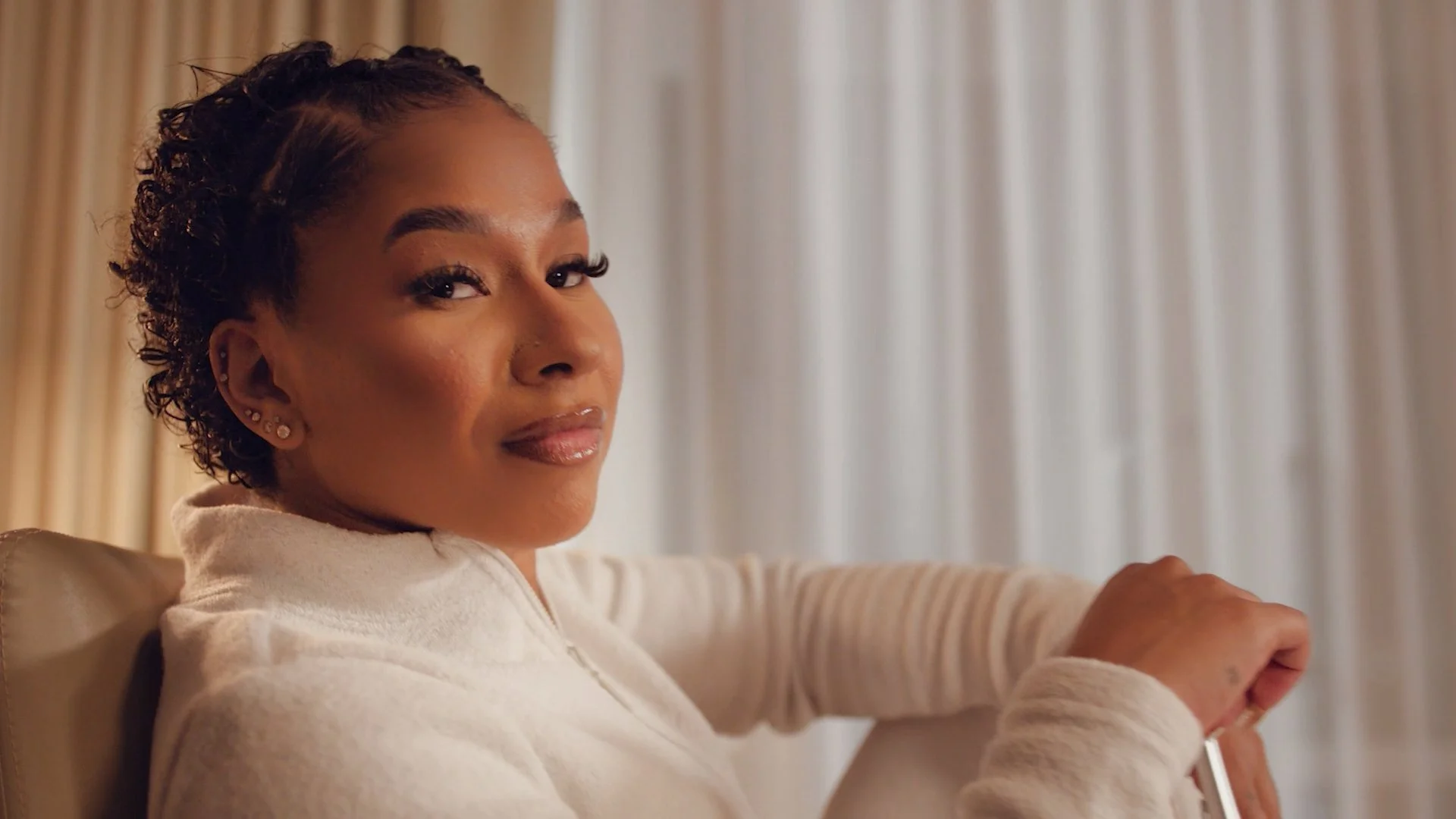 A woman with short, curly hair and multiple earrings, sitting on a beige chair, looking at the camera with a slight smile in front of cream-colored curtains.