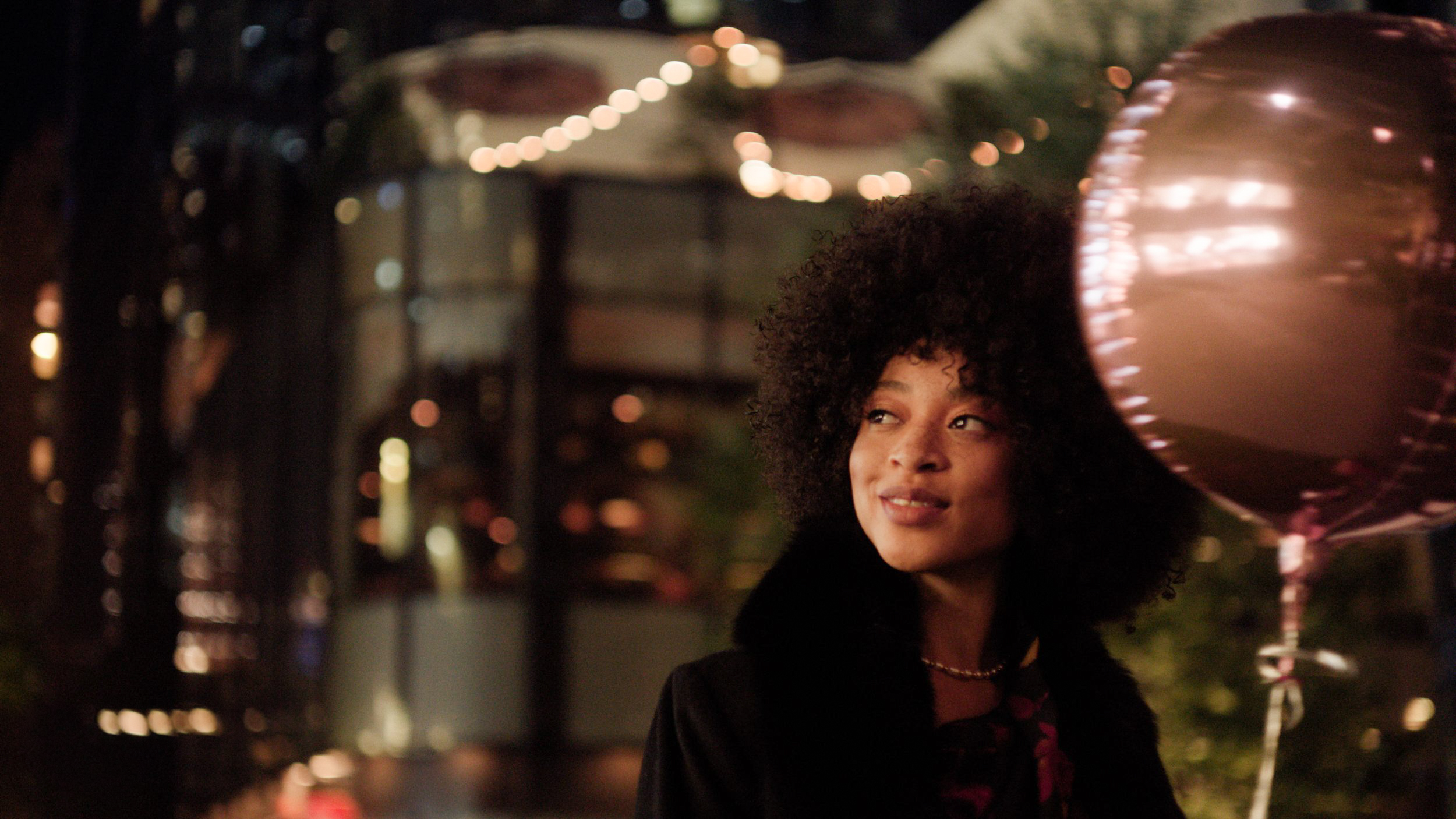A woman with curly hair at night, holding a pink balloon, standing outdoors with blurry string lights in the background.