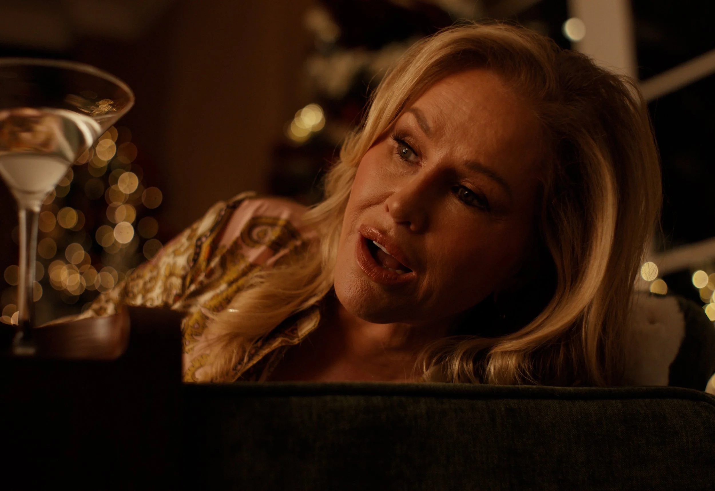 A woman with blonde hair tilting her head, with a surprised or shocked expression, at a party or celebration with blurred lights in the background and a cocktail glass nearby.