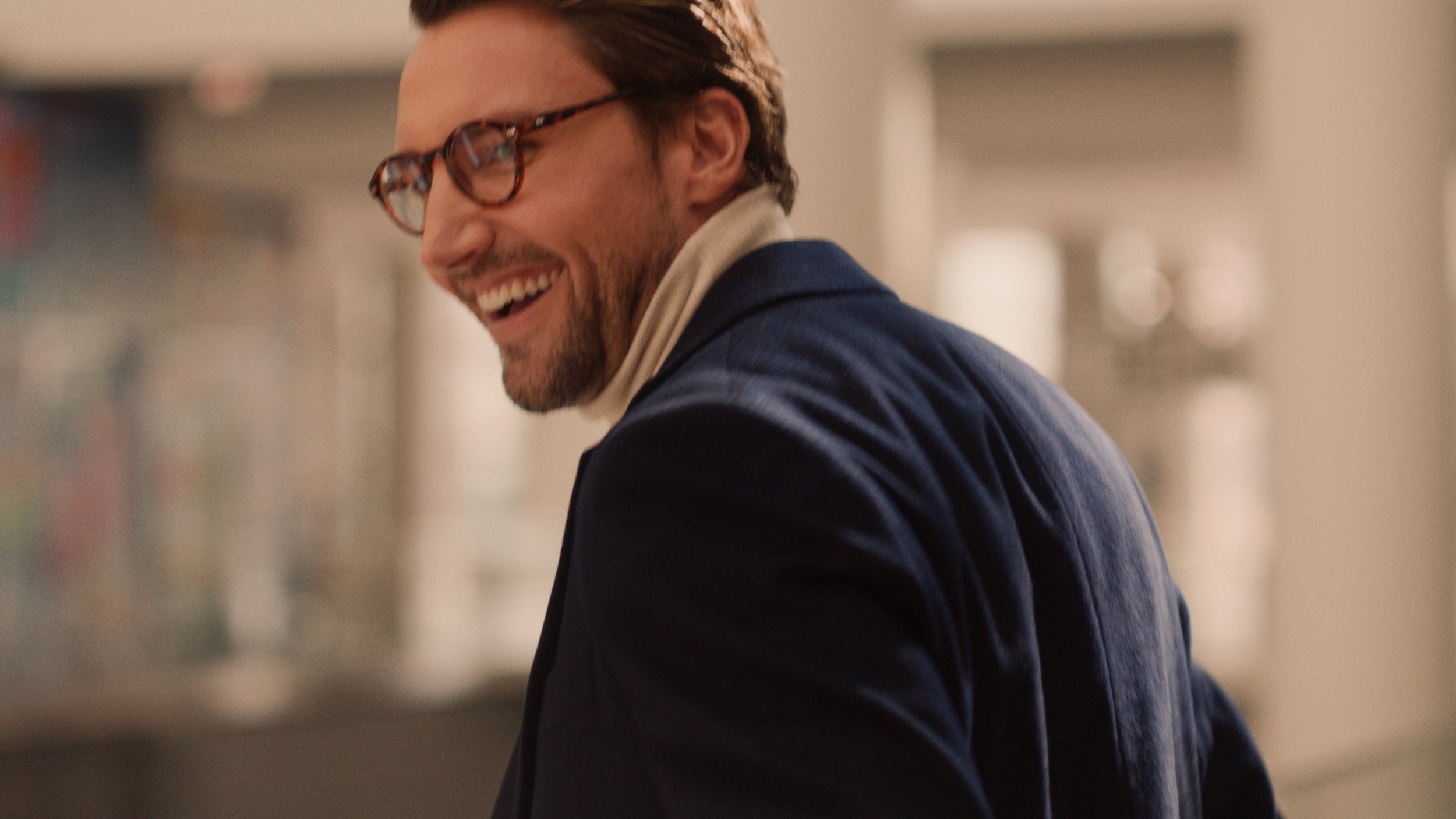 Smiling man with glasses and beard, wearing a navy blue blazer and cream-colored turtleneck, looking to the side in an indoor setting.