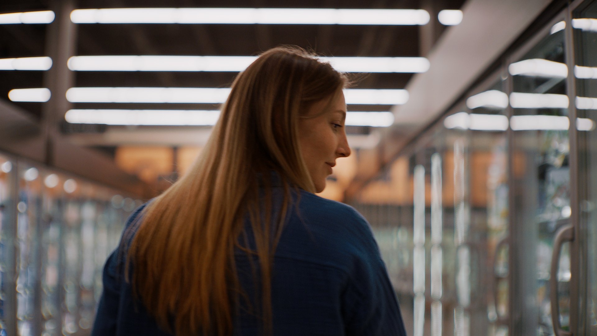 A woman with long red hair and a blue jacket looking to her left inside a store with metal shelves and bright overhead lighting.