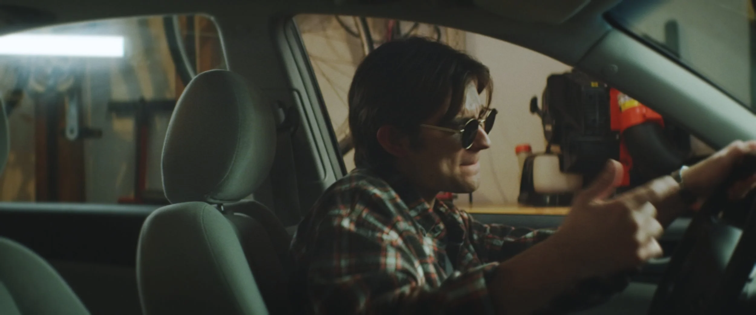 A man with dark hair, wearing sunglasses and a plaid shirt, sitting in the driver's seat of a car, adjusting or holding a camera mounted on the dashboard.