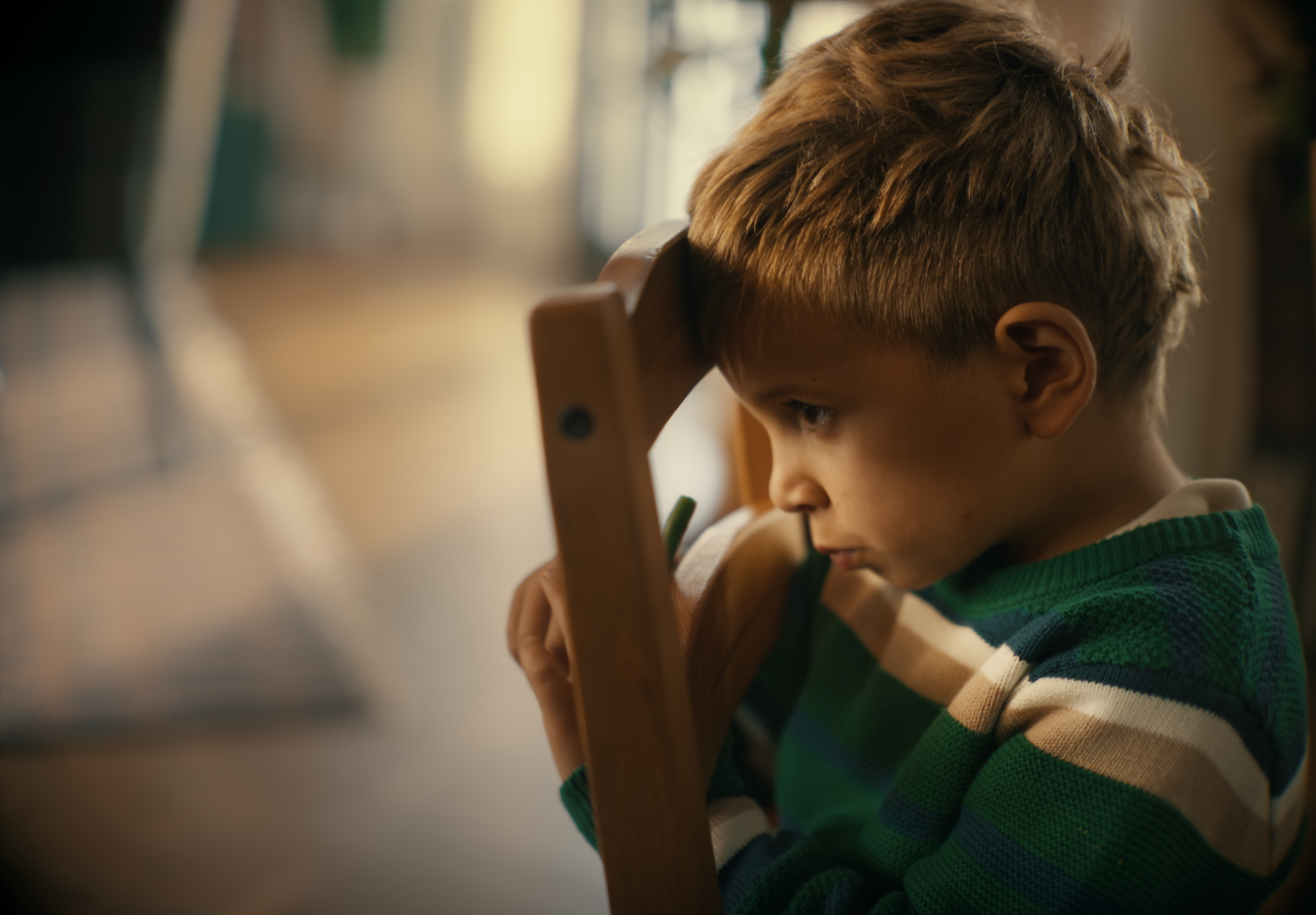 A young boy with light brown hair and a green and white striped sweater is sitting on a wooden chair, resting his head on the back of the chair and looking concerned or deep in thought.