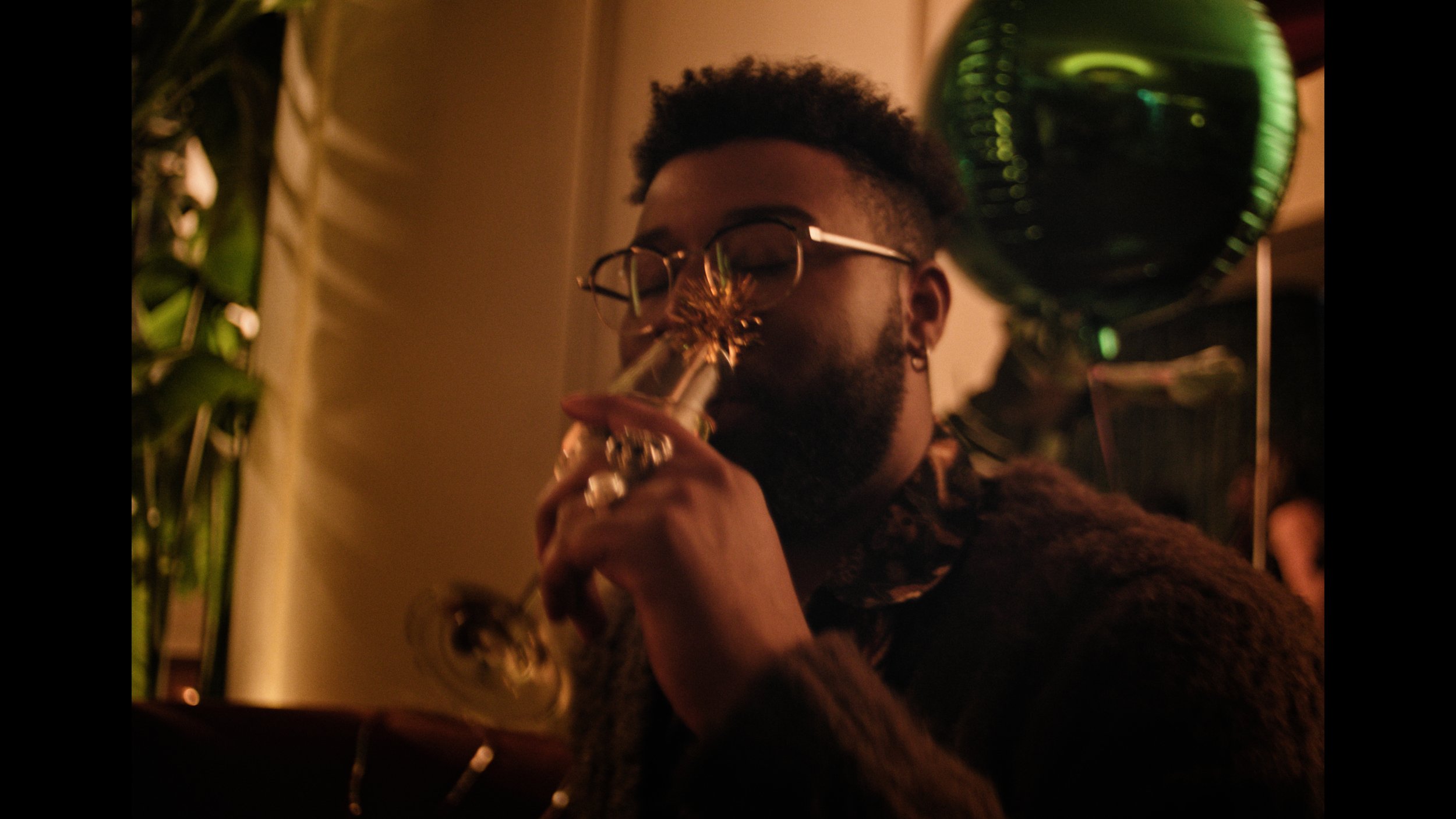 Person with glasses and a beard holding a champagne flute decorated with a star-shaped ornament, with green balloons in the background at a celebration or party.