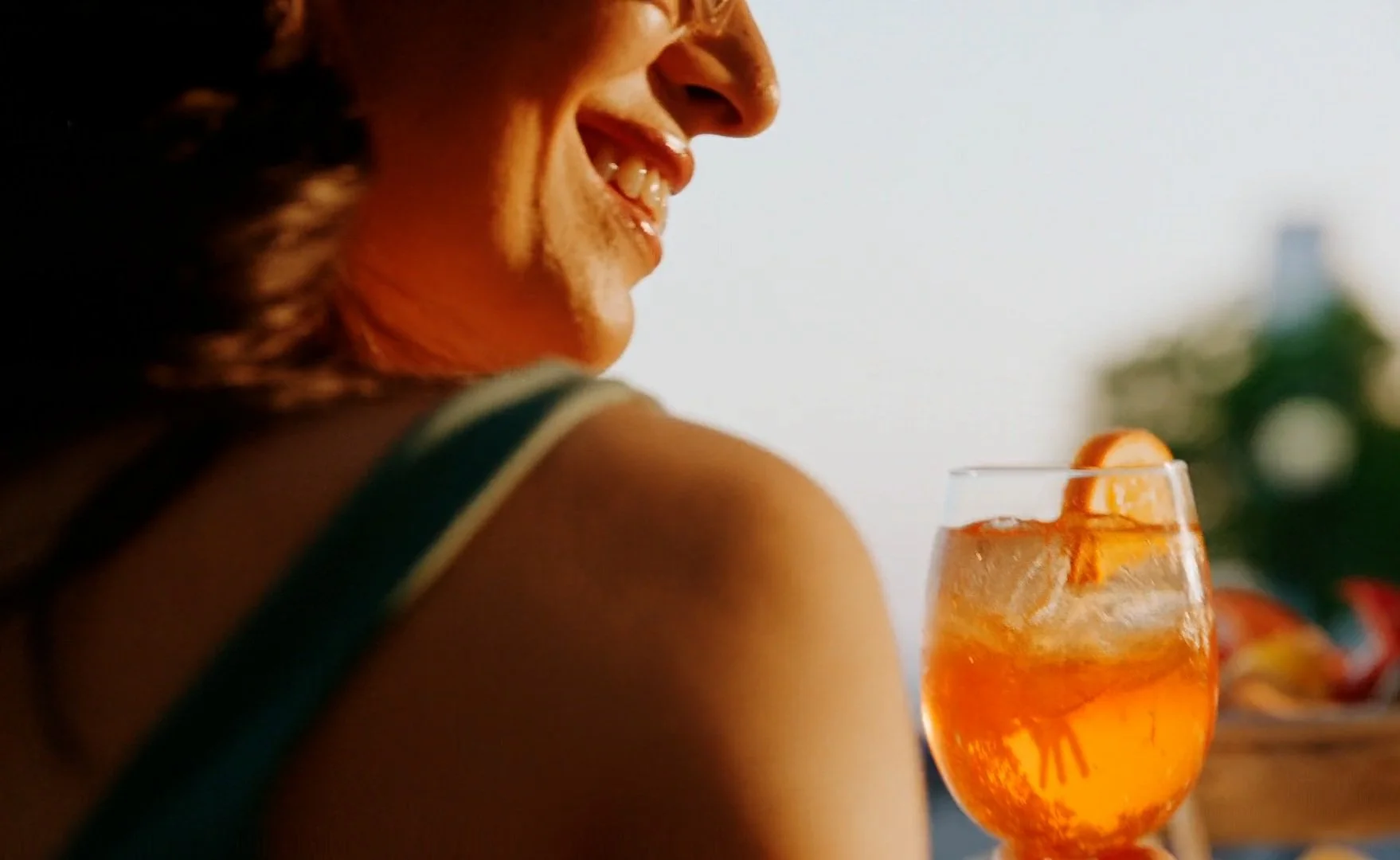 Close-up of a smiling woman looking away, with a glass of orange-colored drink with ice and orange slices in the foreground.