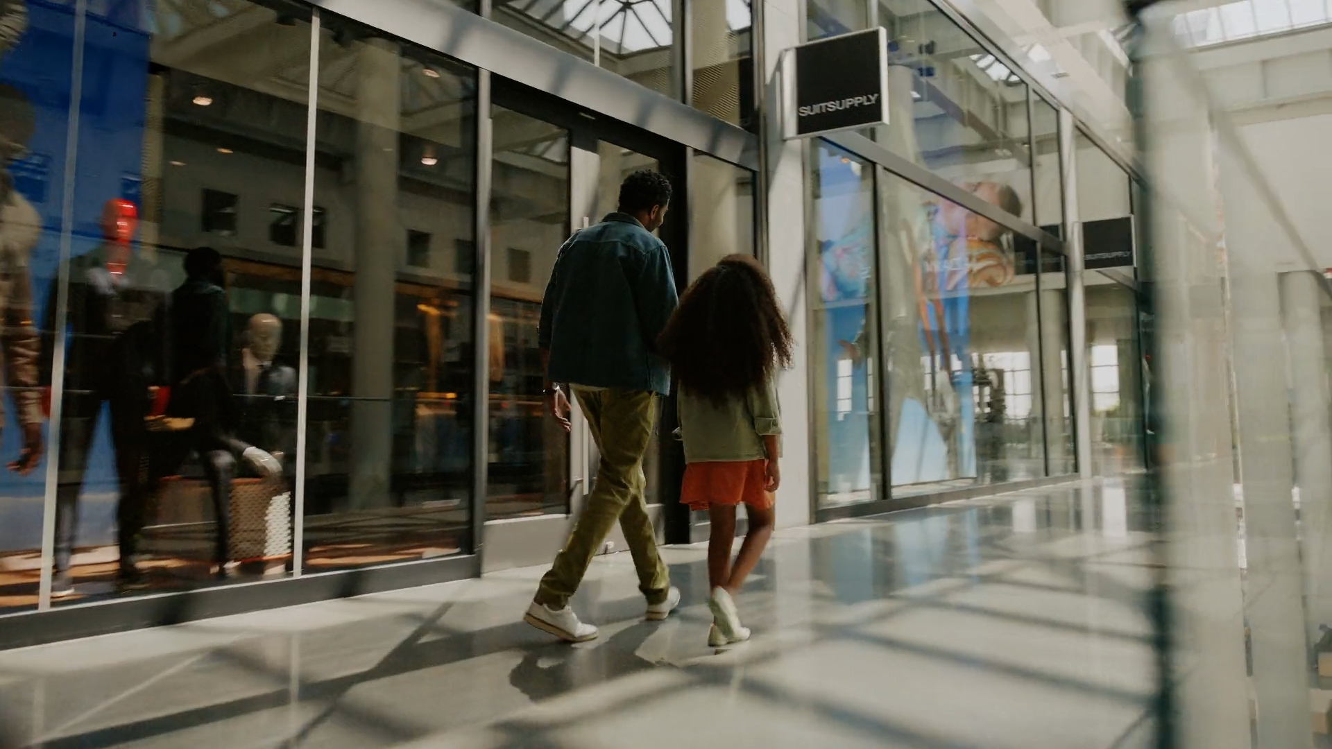 A man and a young girl walking into a shopping mall through glass doors, with reflections of people and store displays on the glass.
